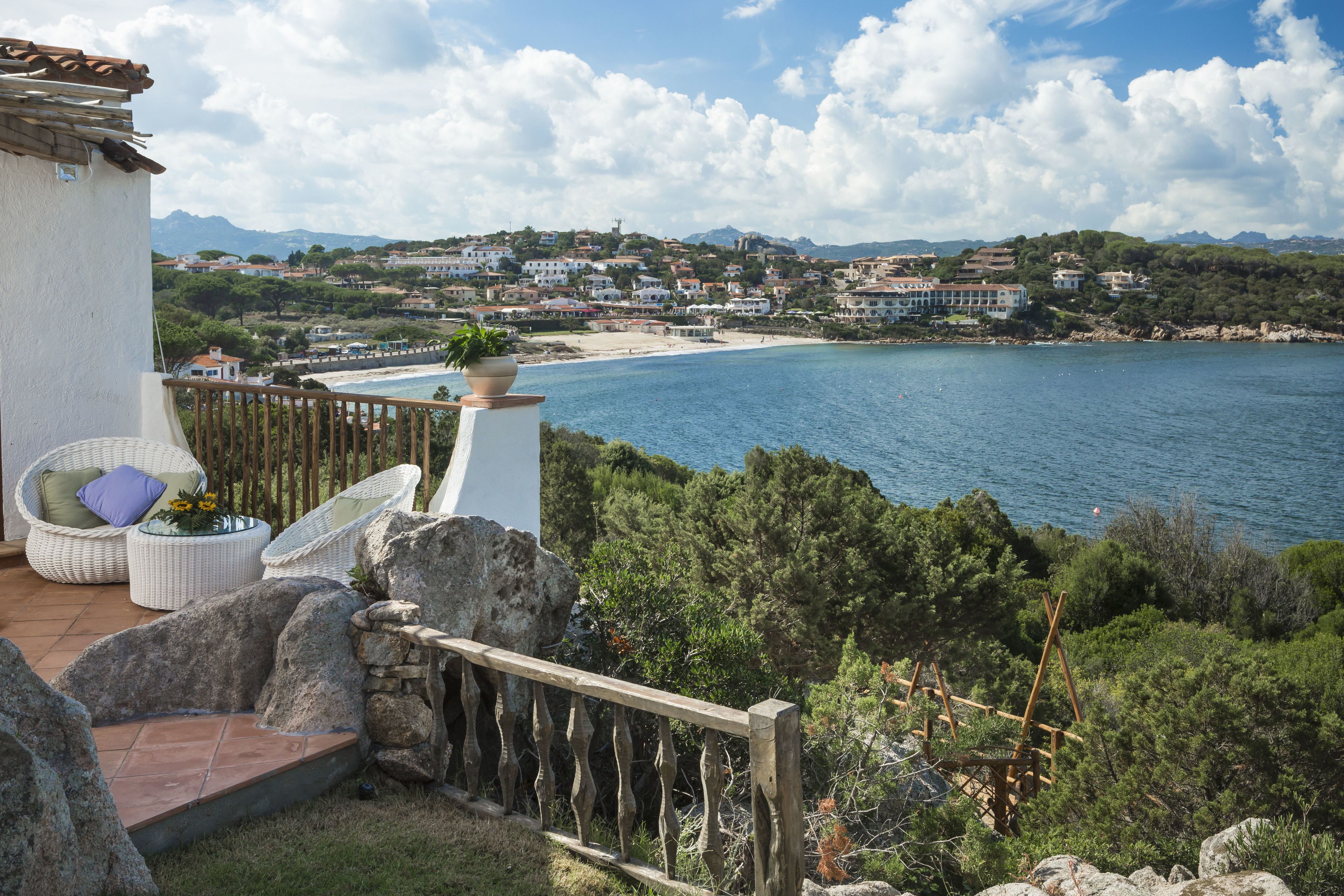 A balcony with wicker furniture, a stone wall, and a view of a coastal town and beach.