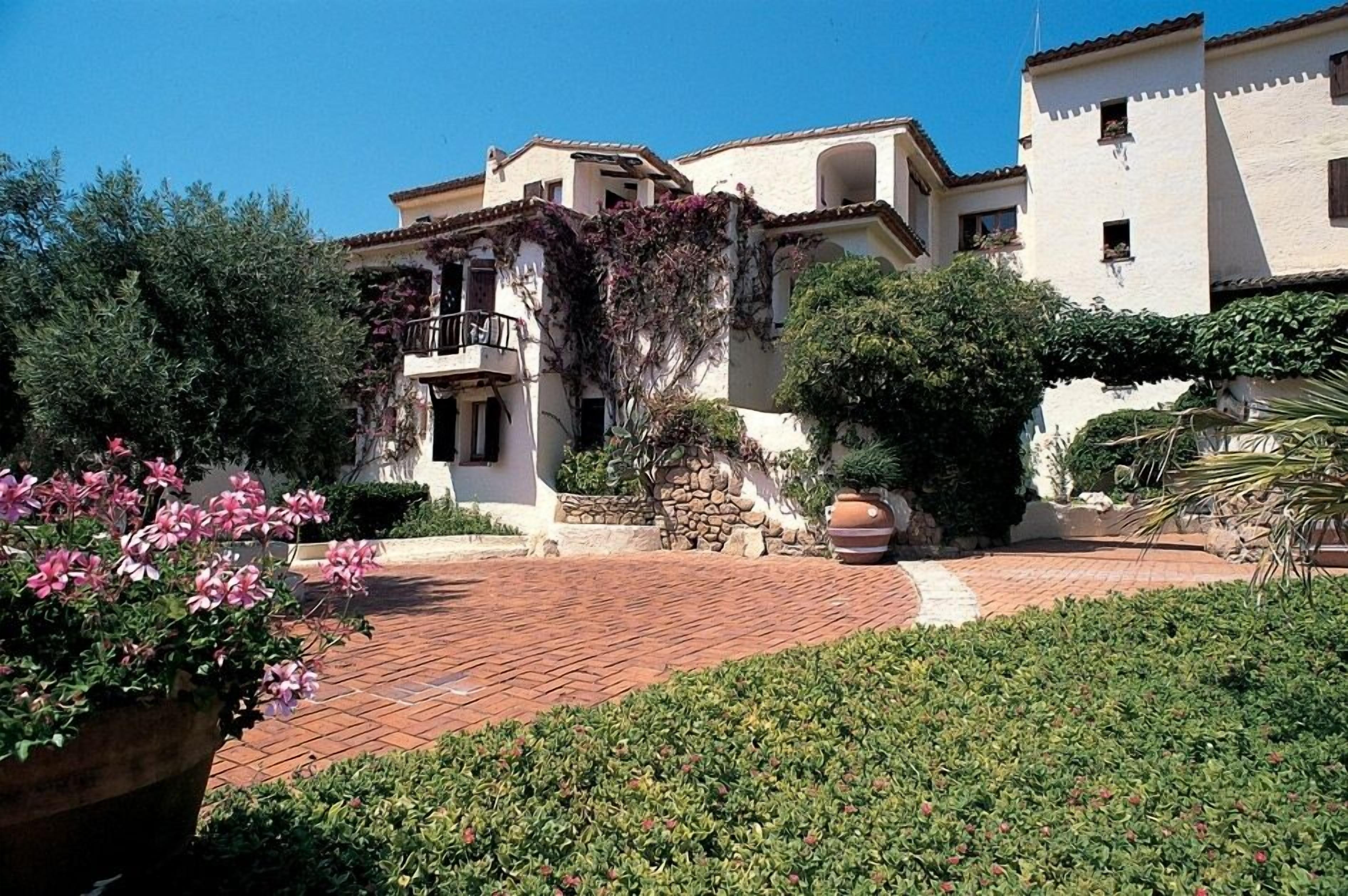 A villa with a paved courtyard, surrounded by lush greenery and a stone wall.