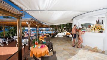 Un espace bar extérieur avec des tables et des chaises, une piscine en arrière-plan et un sol recouvert de carreaux de pierre.