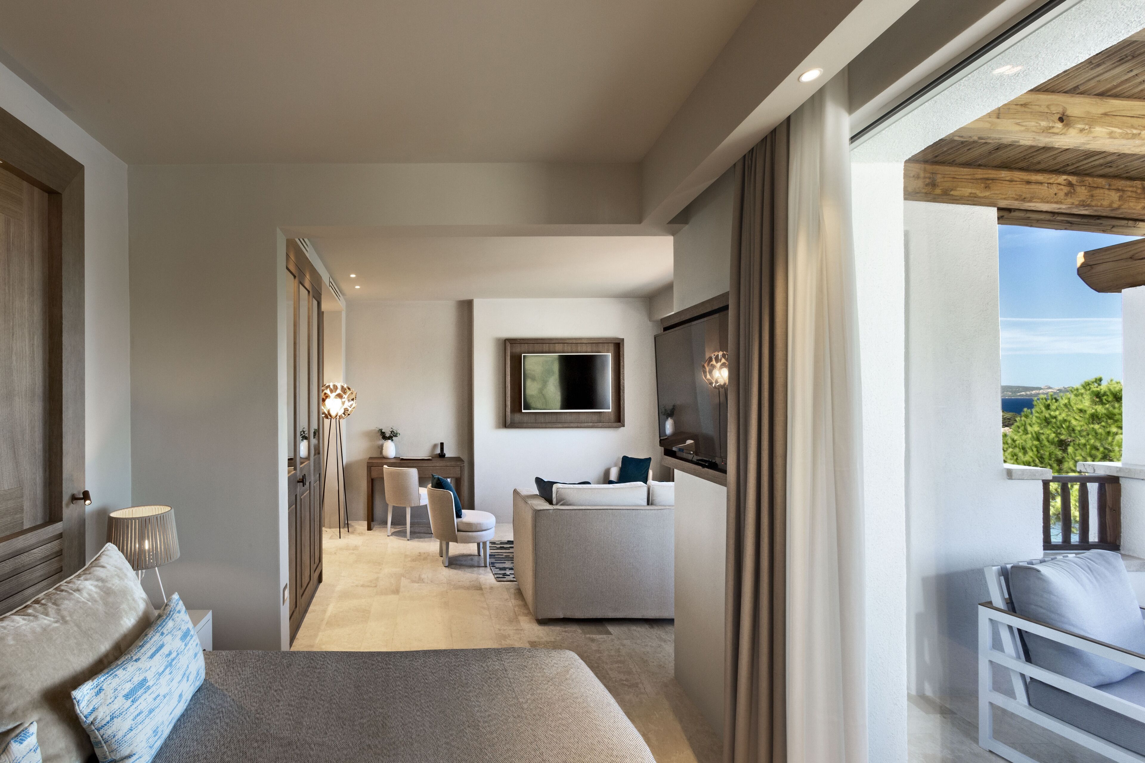 A modern hotel room with a large bed, a seating area with a sofa and chairs, a TV mounted on the wall, and a balcony with a view.