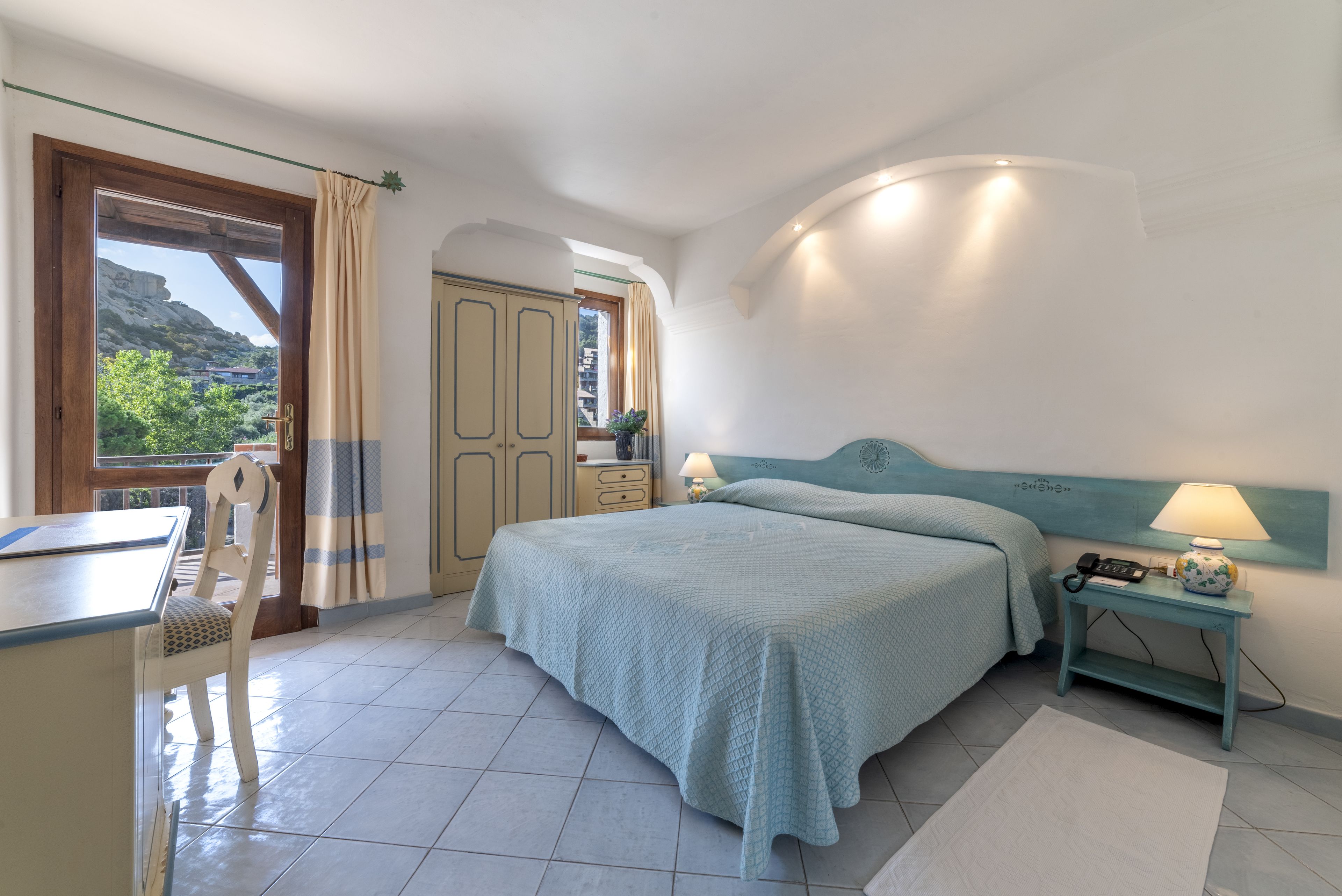 Un dormitorio con cama, mesita de noche, lámpara y vistas al exterior.