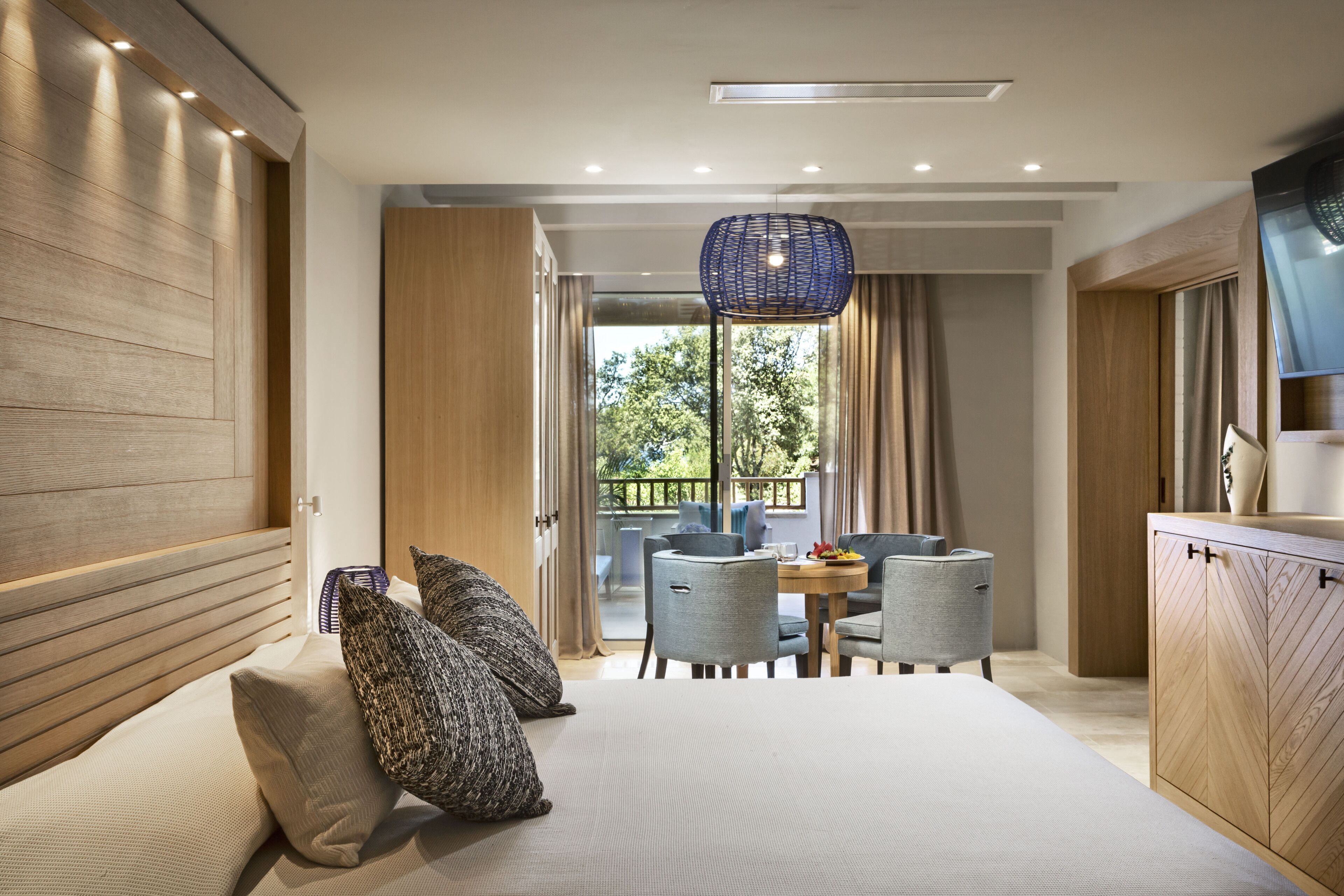 A modern hotel room with a large bed, a dining area with chairs, a television, and a view of the outdoors.