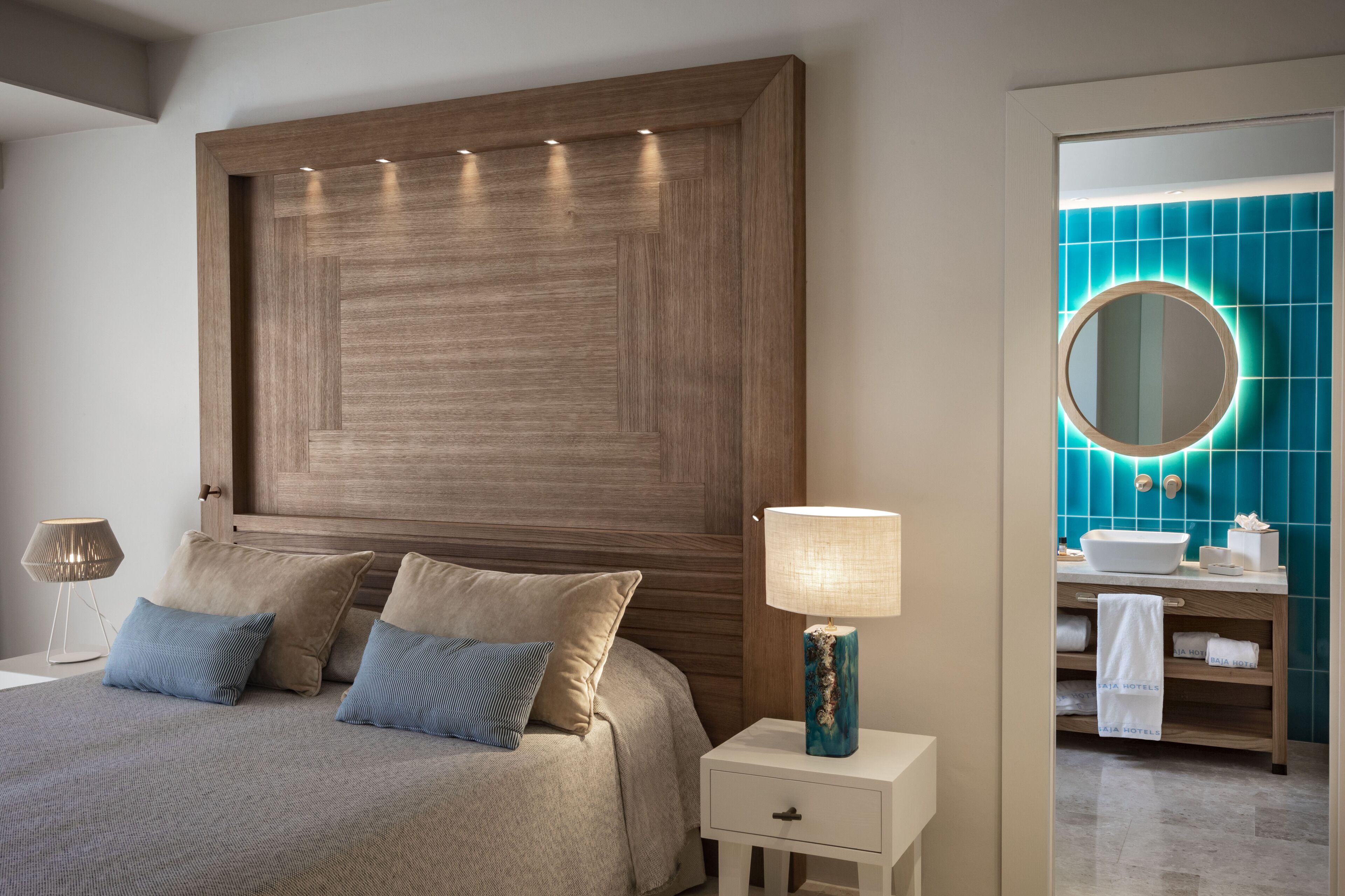 Een moderne slaapkamer met een houten hoofdbord, een nachtkastje met een lamp, en uitzicht op een badkamer met een blauwe betegelde muur en een ronde spiegel.