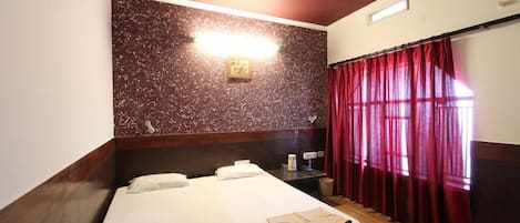 Une chambre d’hôtel avec un lit, une table de chevet, une lampe, une horloge murale et une fenêtre avec des rideaux rouges.