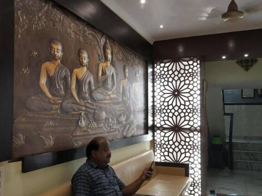 A man sitting on a bench in a room with a large Buddha mural on the wall, a decorative partition, and a ceiling fan.