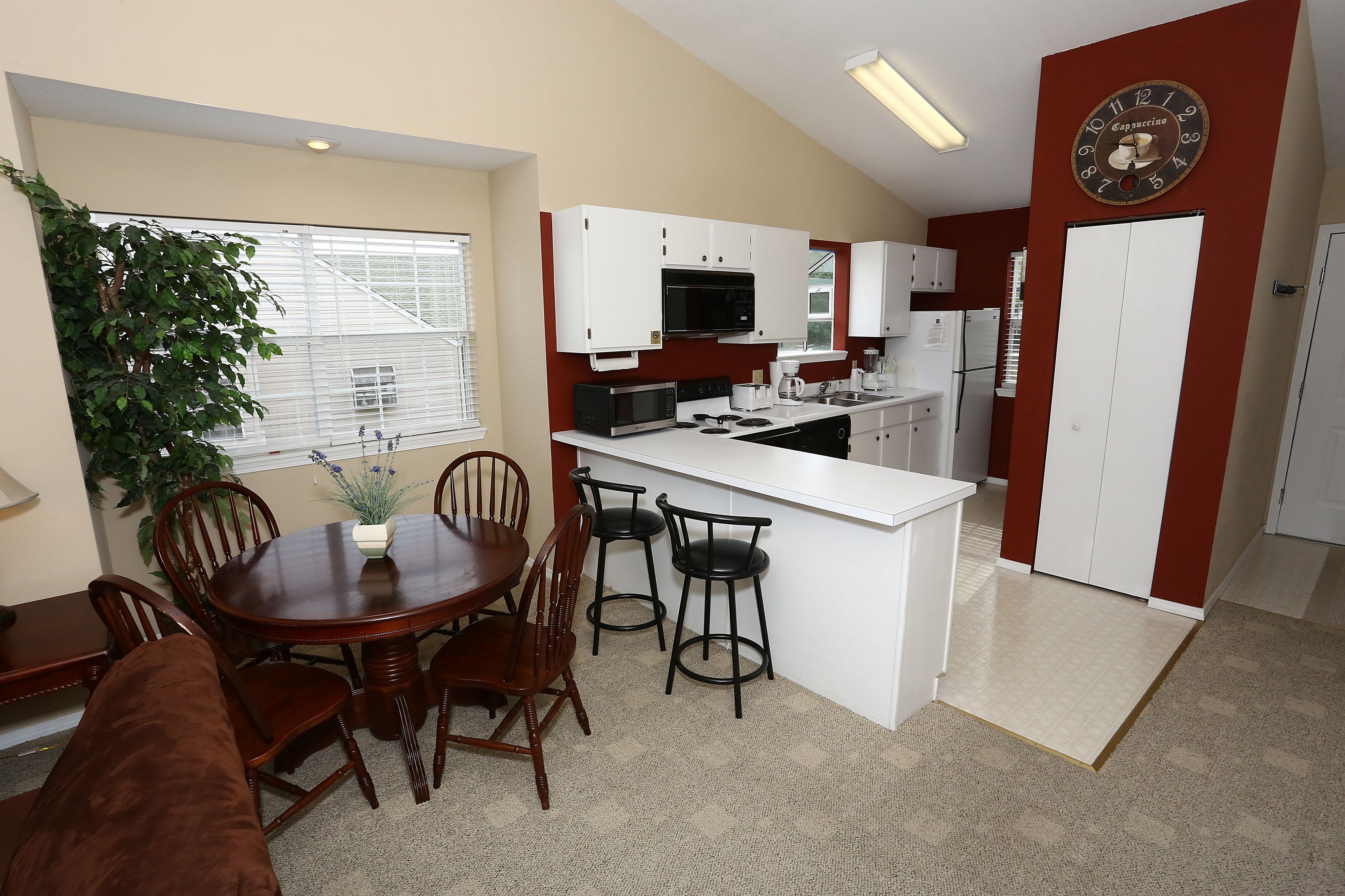 A compact kitchen with white cabinets, a microwave, and a coffee maker. There is a dining area with a wooden table and chairs.