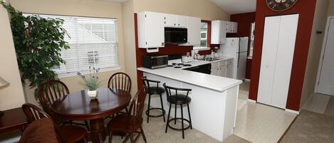 A compact kitchen with white cabinets, a microwave, and a coffee maker. There is a dining area with a wooden table and chairs.