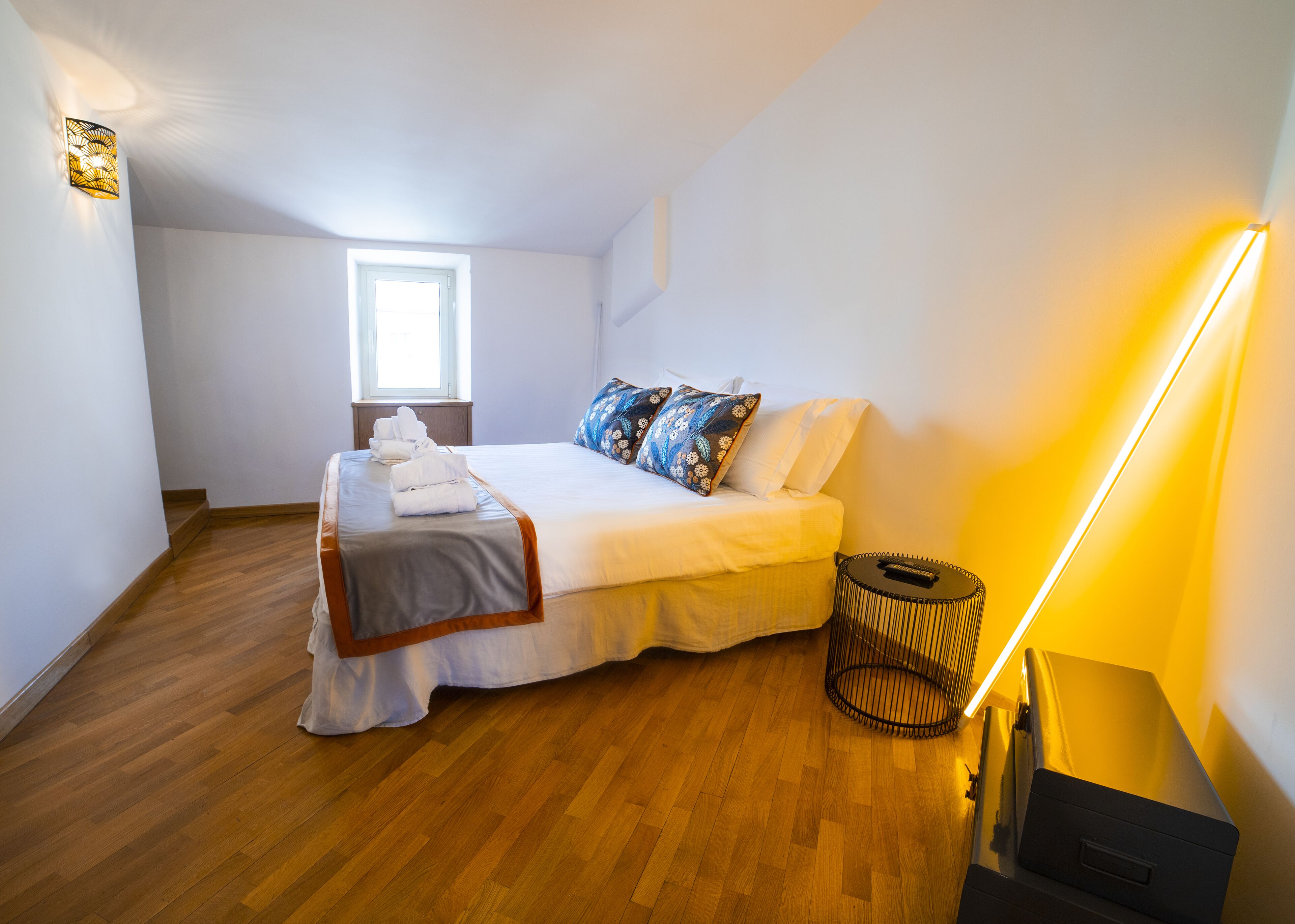 A neatly made bed with a grey bedspread and white linens, a window with natural light, a bedside lamp, and a small round table with a lamp.