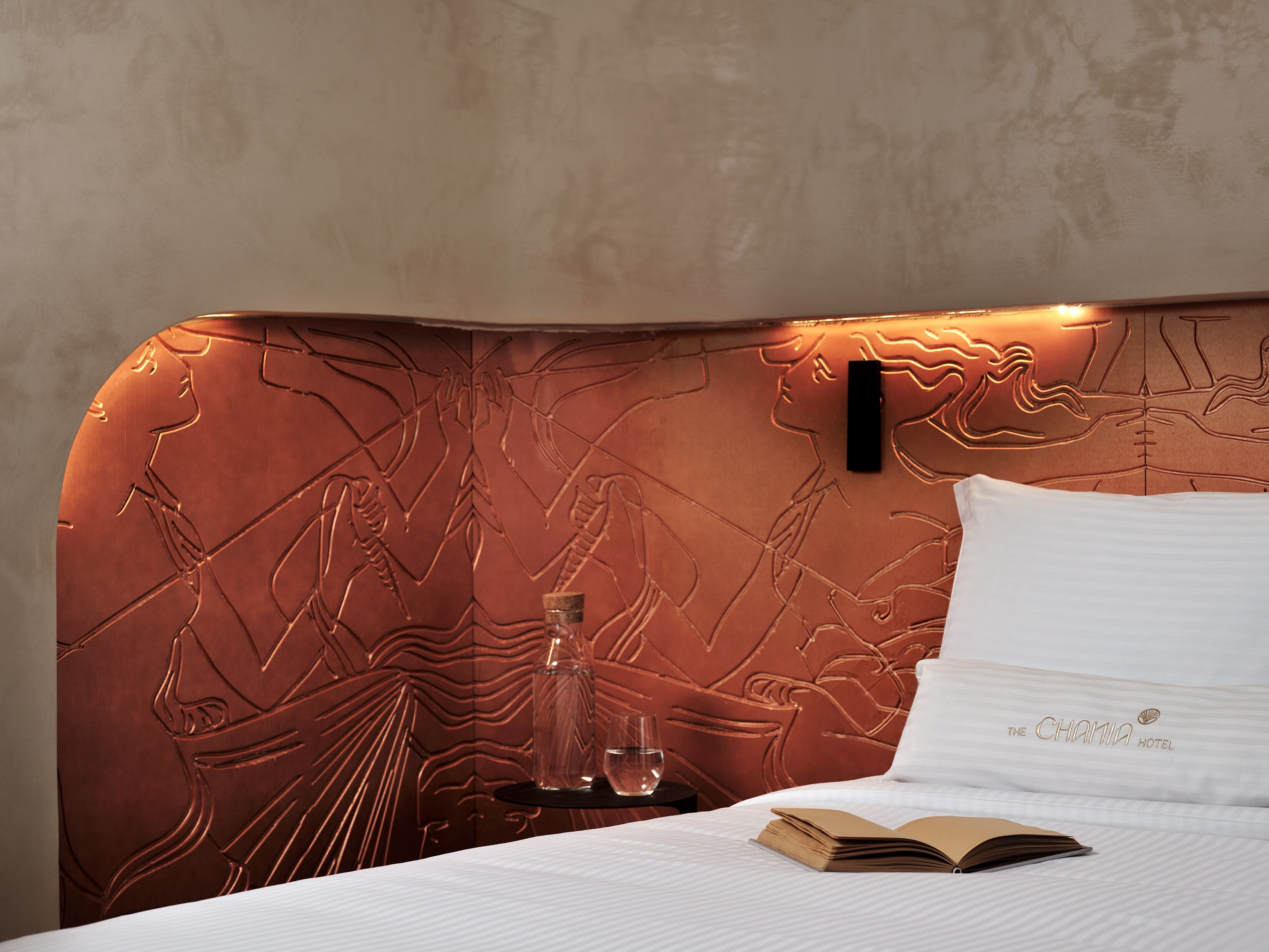 Une chambre d’hôtel avec une tête de lit à motifs, une table de chevet sur laquelle se trouve un verre d’eau et un livre posé sur le lit.