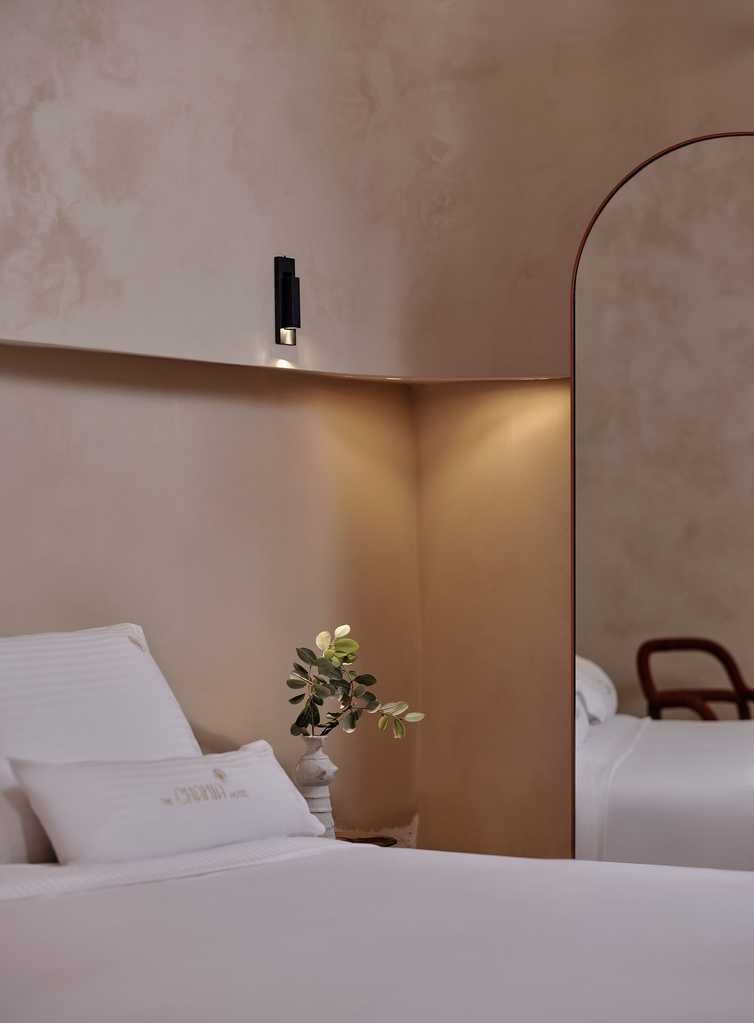 Una habitación de hotel con una cama, una mesita de noche con un florero, una lámpara de pared y un espejo grande.