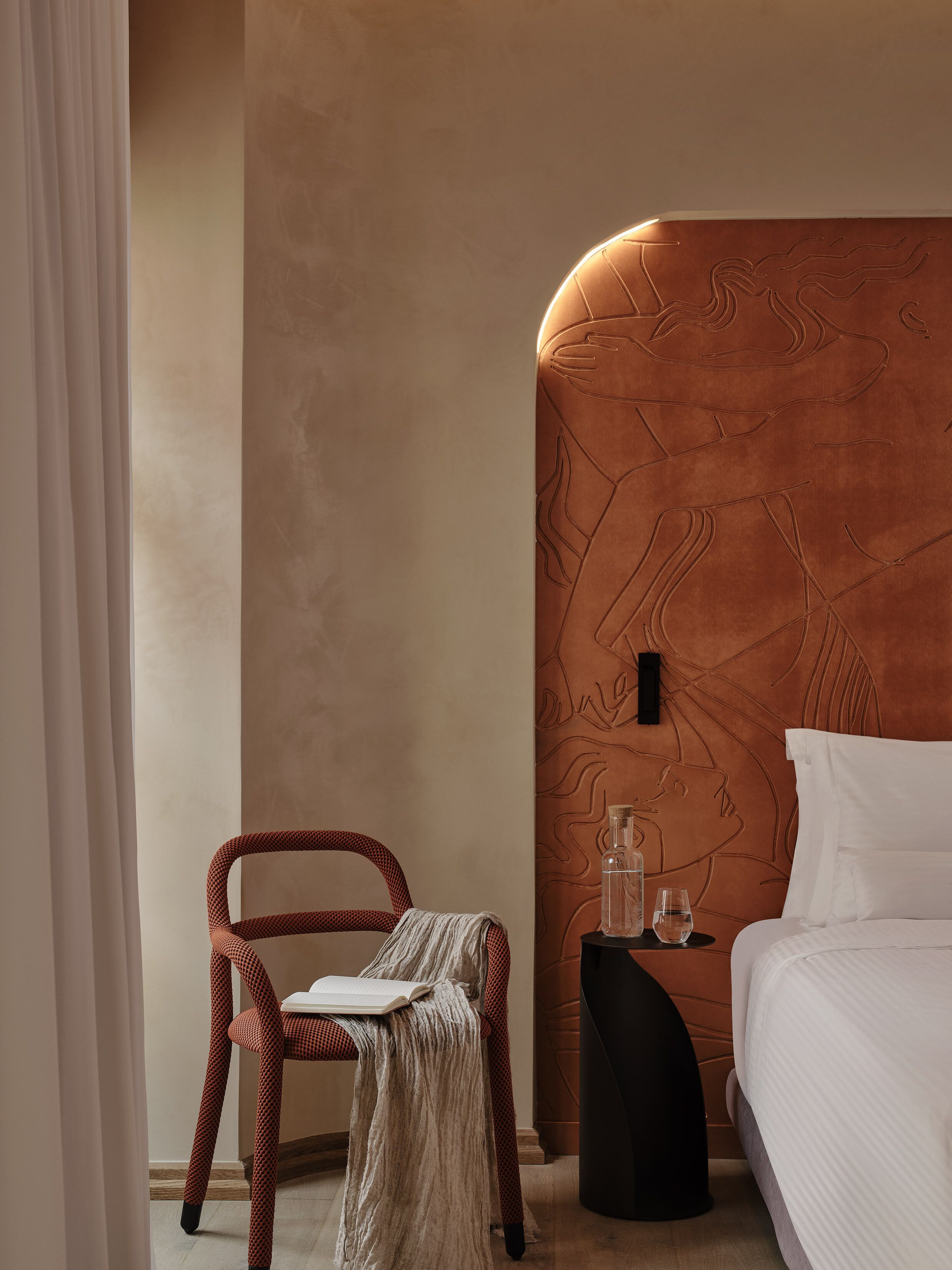 A bedroom with a headboard, a bedside table with a bottle and glasses, a chair with a book, and a bed with white bedding.