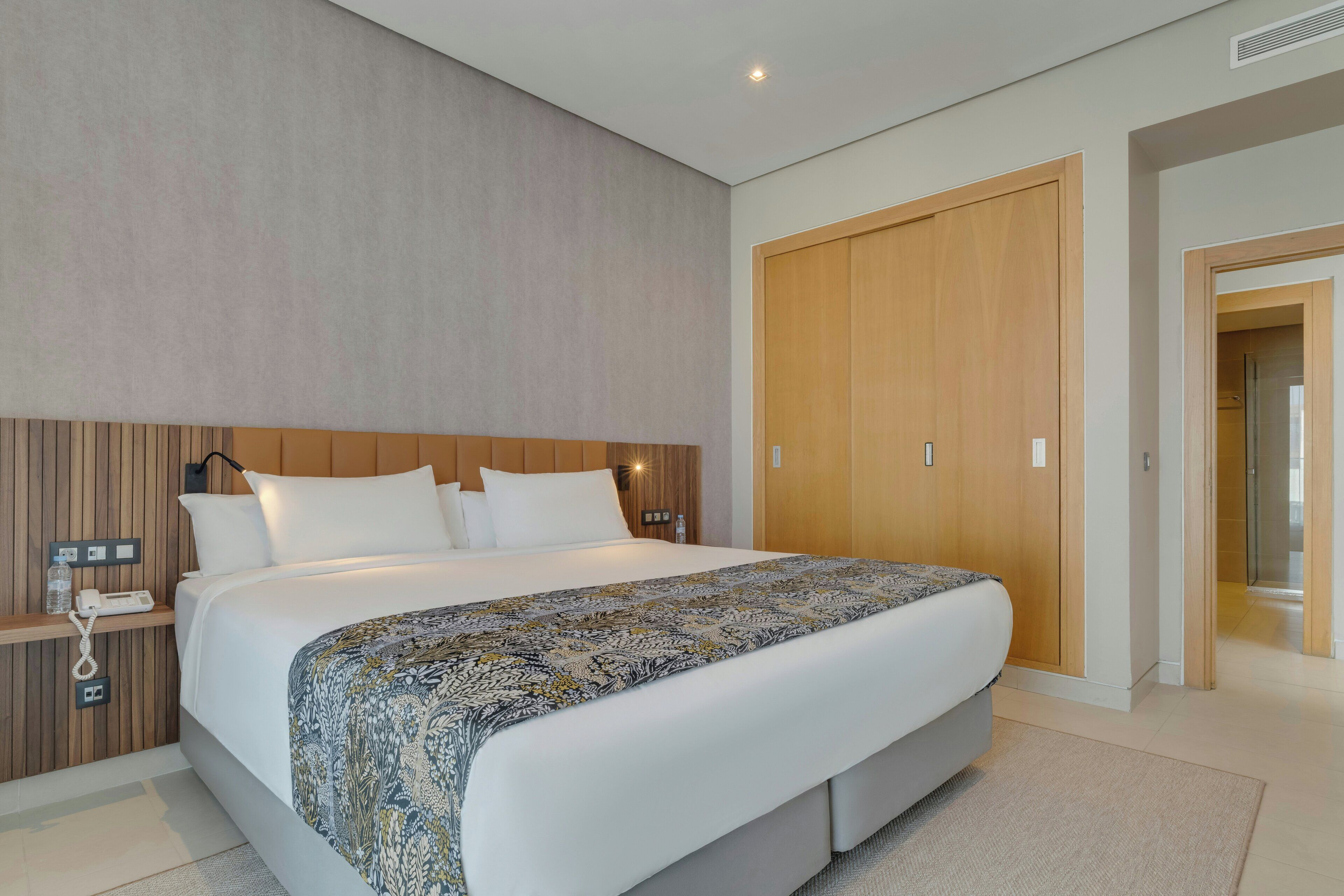Habitación de hotel moderna con una cama grande, cabecera de madera, mesita de noche con un teléfono y una botella de agua, y un armario.