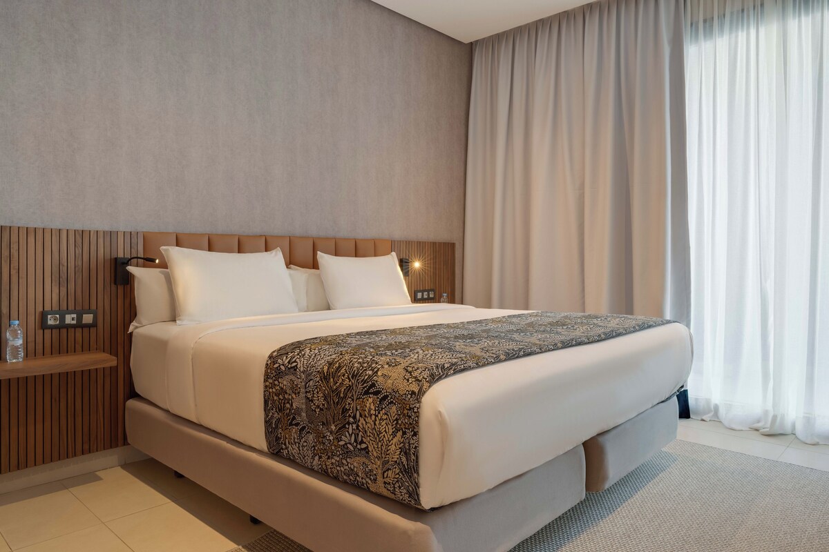 Een moderne hotelkamer met een groot bed, een houten hoofdbord en een nachtkastje met een waterfles.