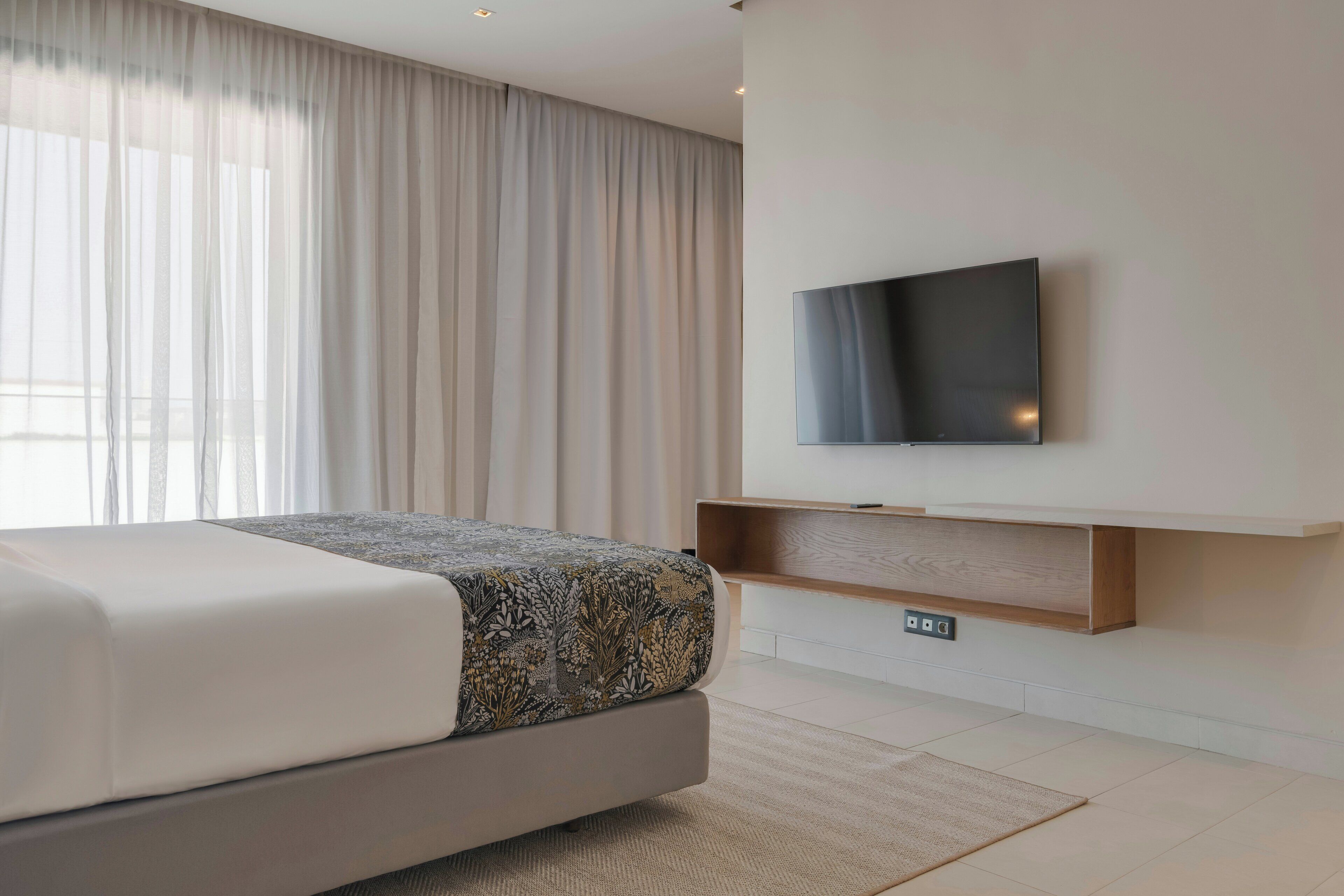 A modern hotel room with a bed, a flat-screen TV mounted on the wall, and a wooden shelf below it.