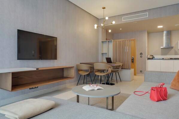 A modern living room with a flat-screen TV, a round coffee table with a book and glasses, a red handbag, and a kitchen area in the background. - Pestana Casablanca (Casablanca)