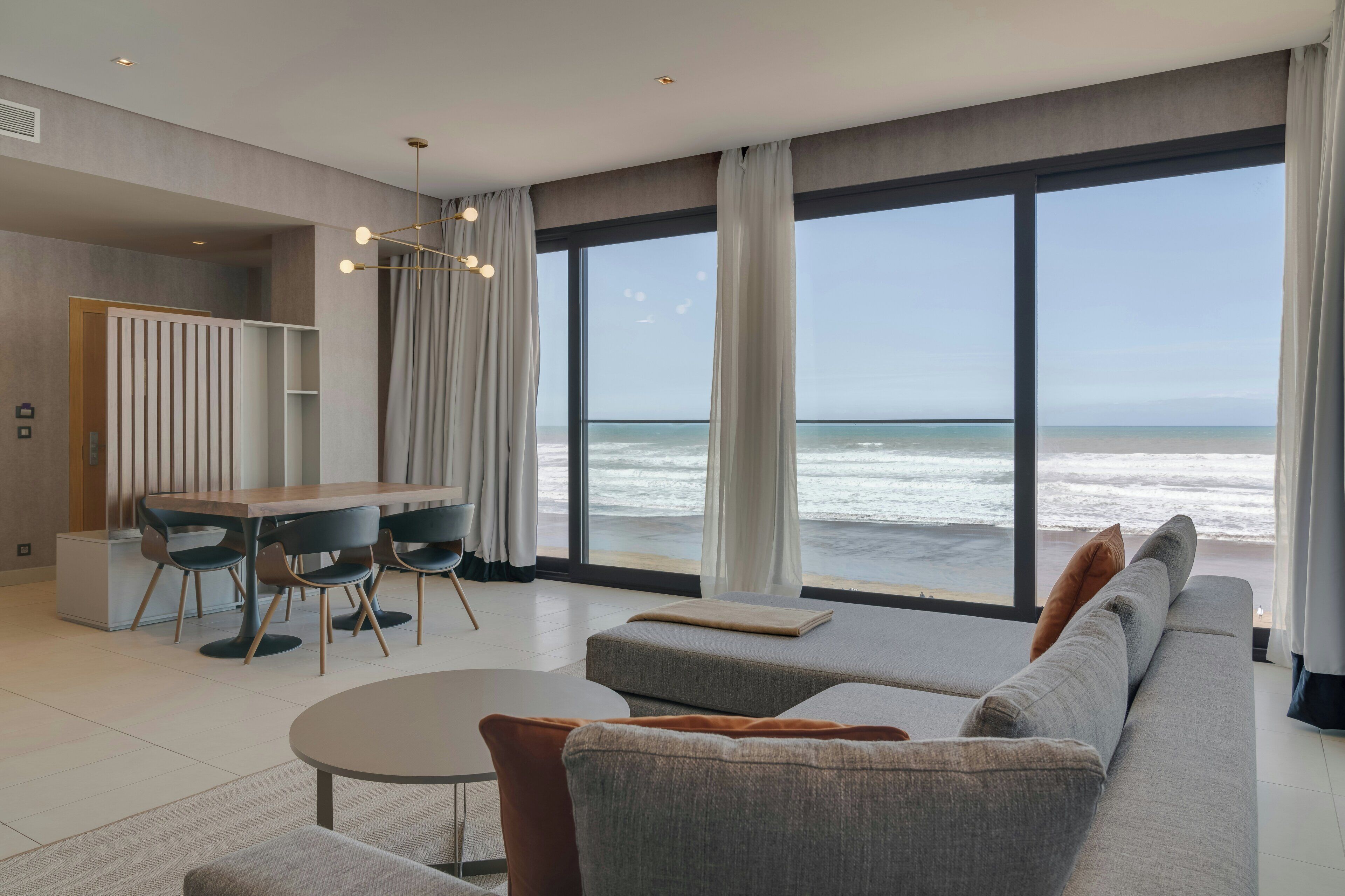 Uma sala de estar moderna com uma janela ampla com vista para o oceano, uma área de jantar com mesa e cadeiras, e um sofá com almofadas.