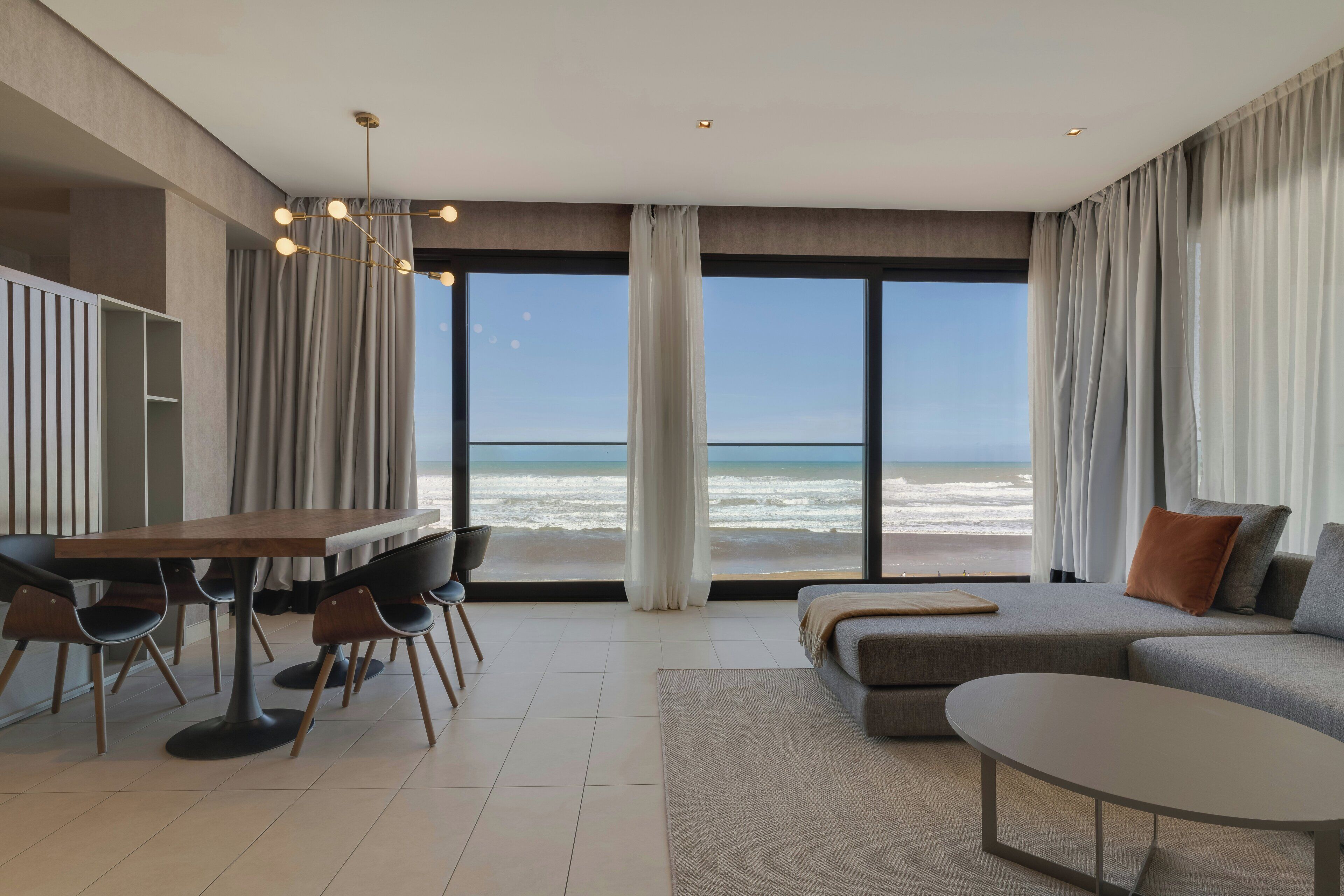 A modern living room with a large window overlooking the beach, a dining area with chairs and a table, and a sofa with cushions.
