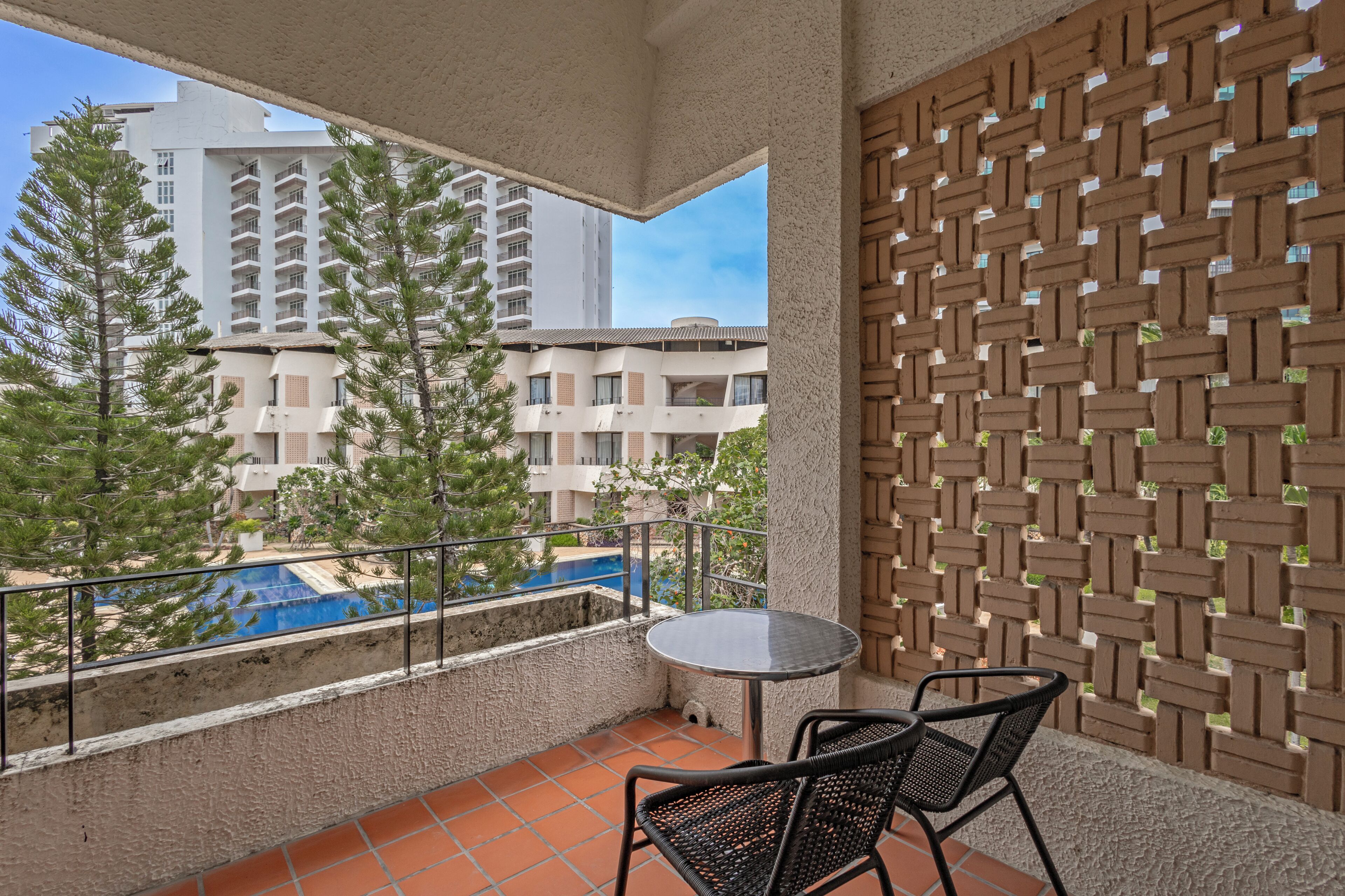 A balcony with a view of a swimming pool, surrounded by tall trees and a brick wall.