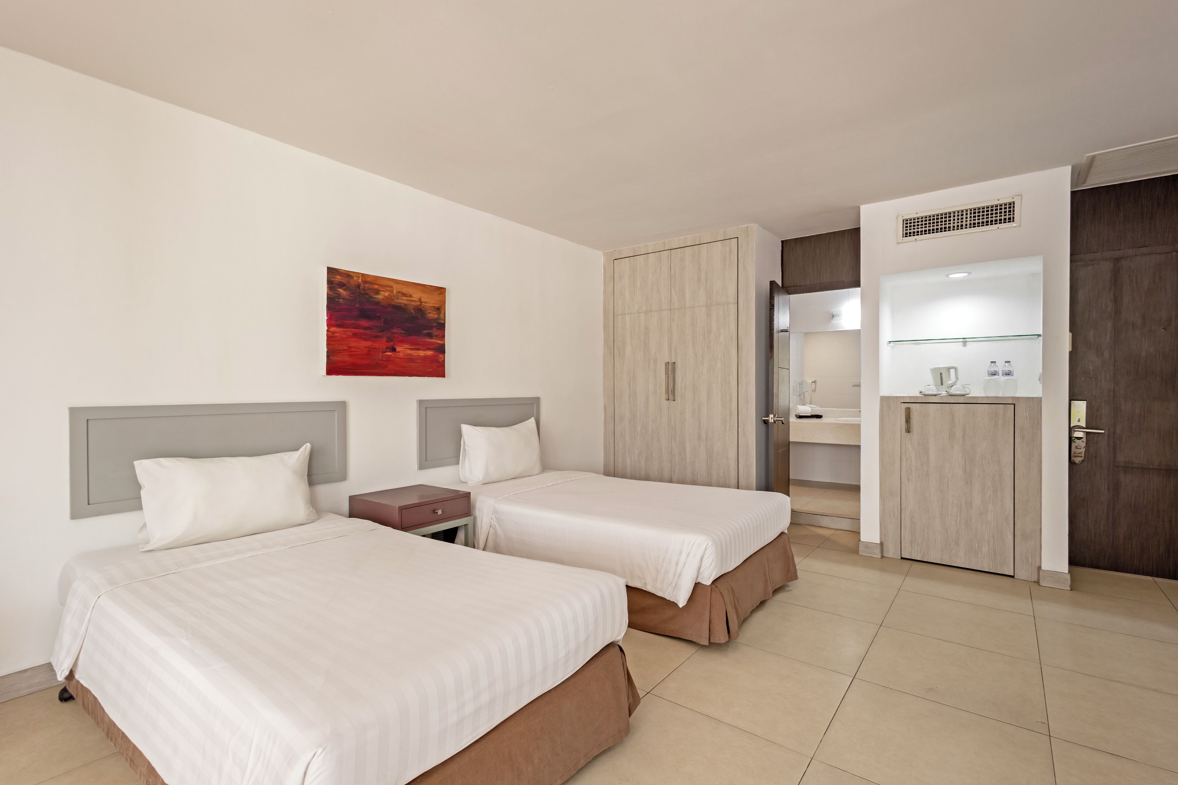 A hotel room with two beds, a painting, a wardrobe, and a kitchenette.