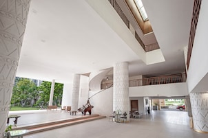 Un vestíbulo amplio con techos altos, grandes columnas y una escalera central. Hay zonas de estar y vistas al exterior.
