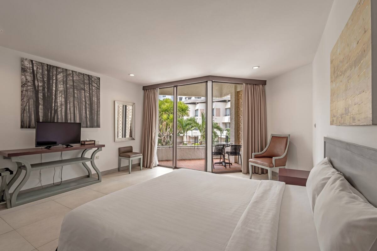 A hotel room with a large bed, a TV on a console table, a desk with a chair, and a balcony with a table and chairs.