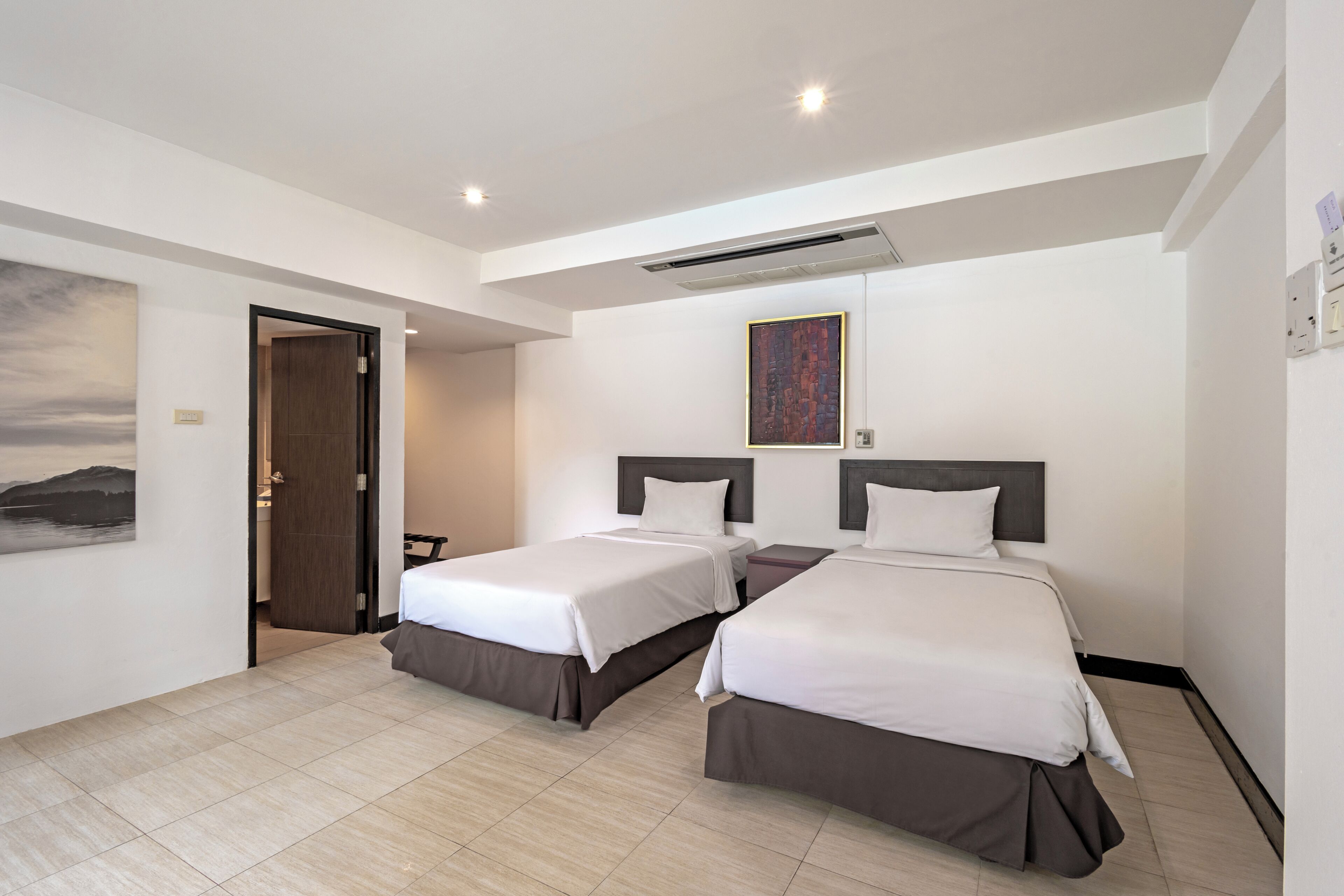 Habitación de hotel con dos camas, un cuadro en la pared y una puerta que da a otra habitación.