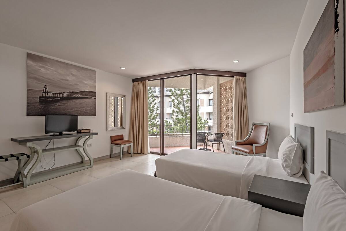 A hotel room with two beds, a TV, a desk, and a balcony with a view of the city.