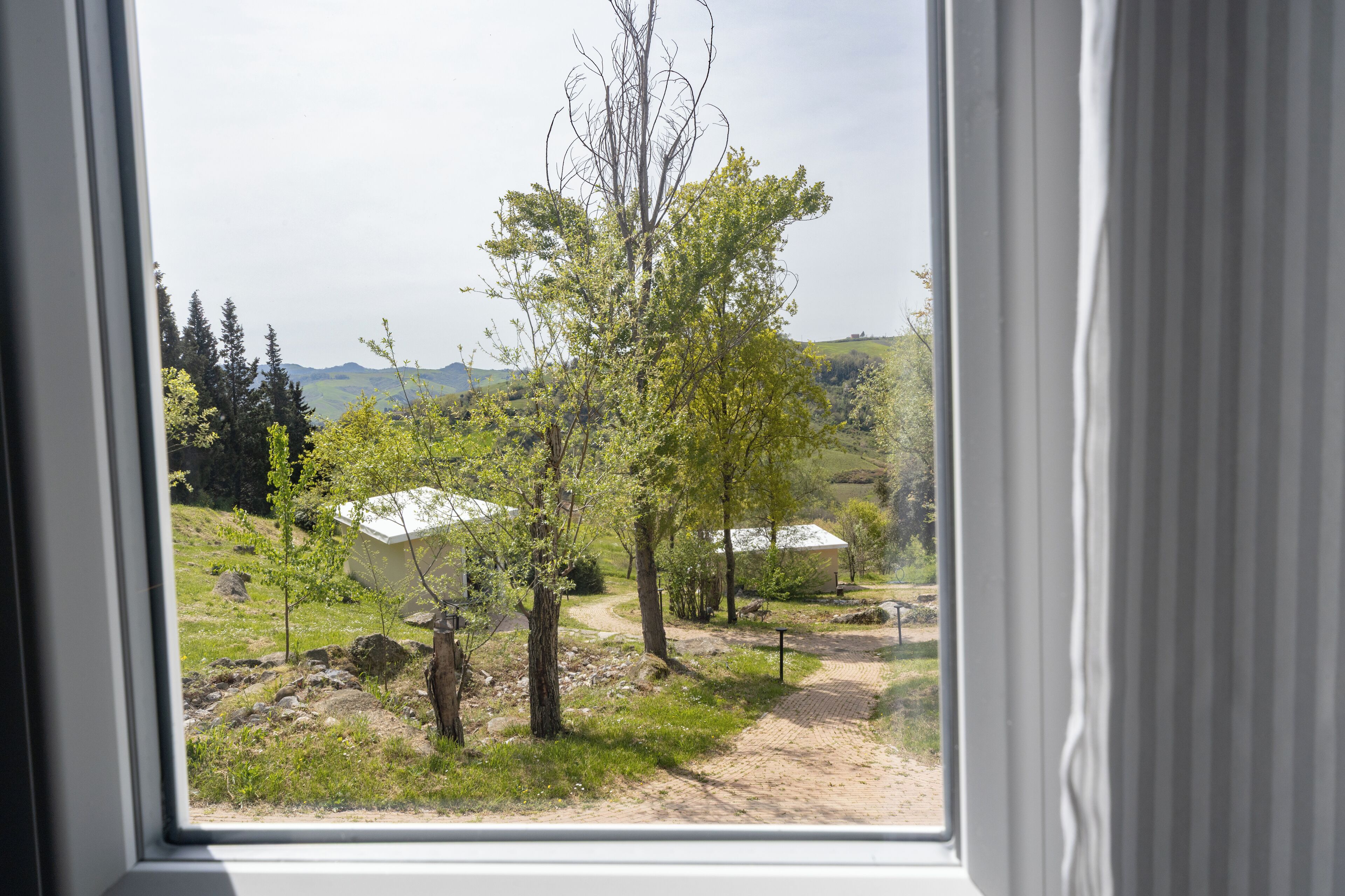 Vista per una finestra d'un paisatge rural amb arbres, un camí de terra i un edifici amb teulada blanca.