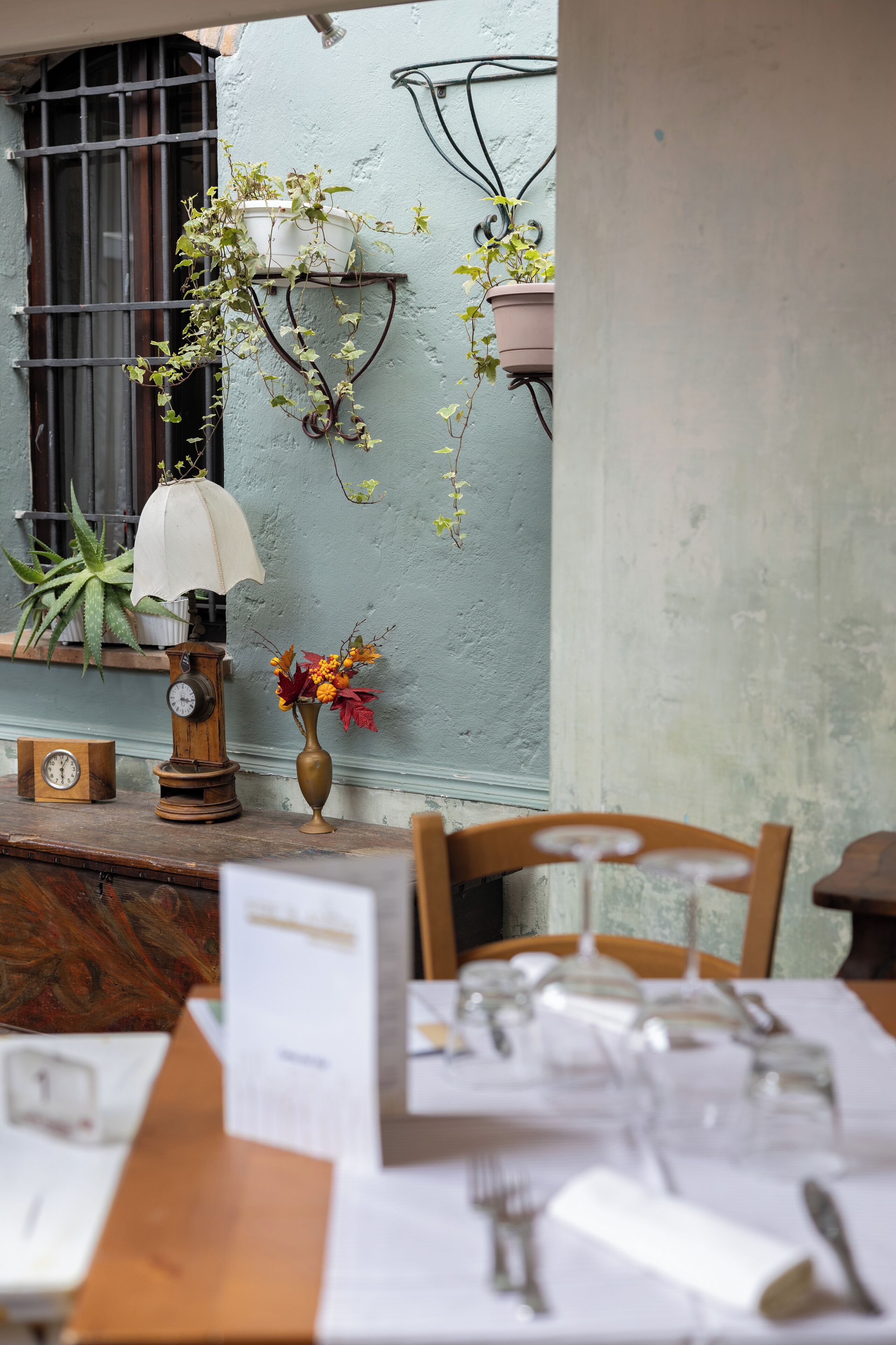 A dining area with a table set for two, a wall-mounted clock, a hanging plant, and a vase with flowers.