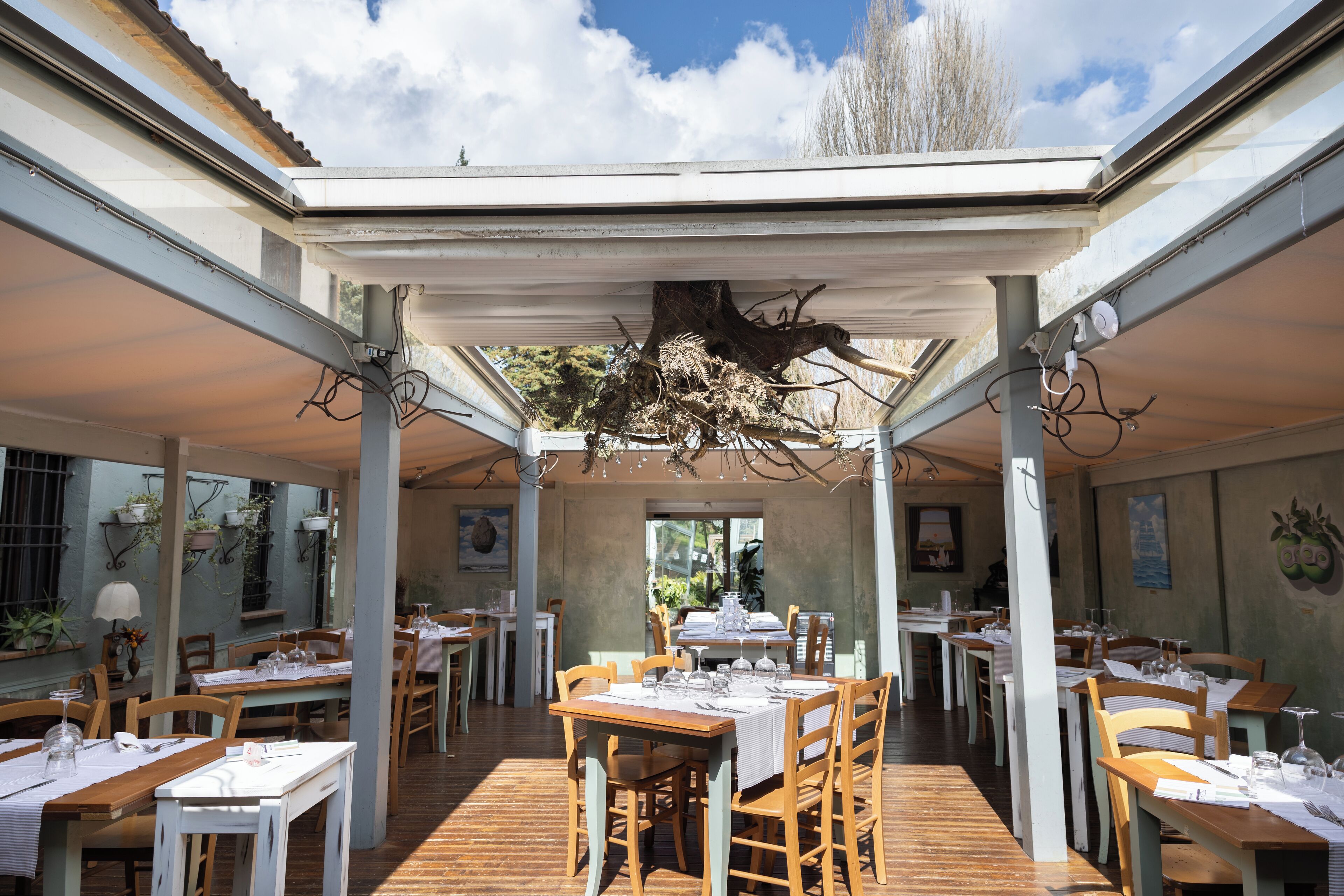 An outdoor dining area with wooden tables and chairs, covered by a pergola, and a bird's nest hanging from the ceiling.