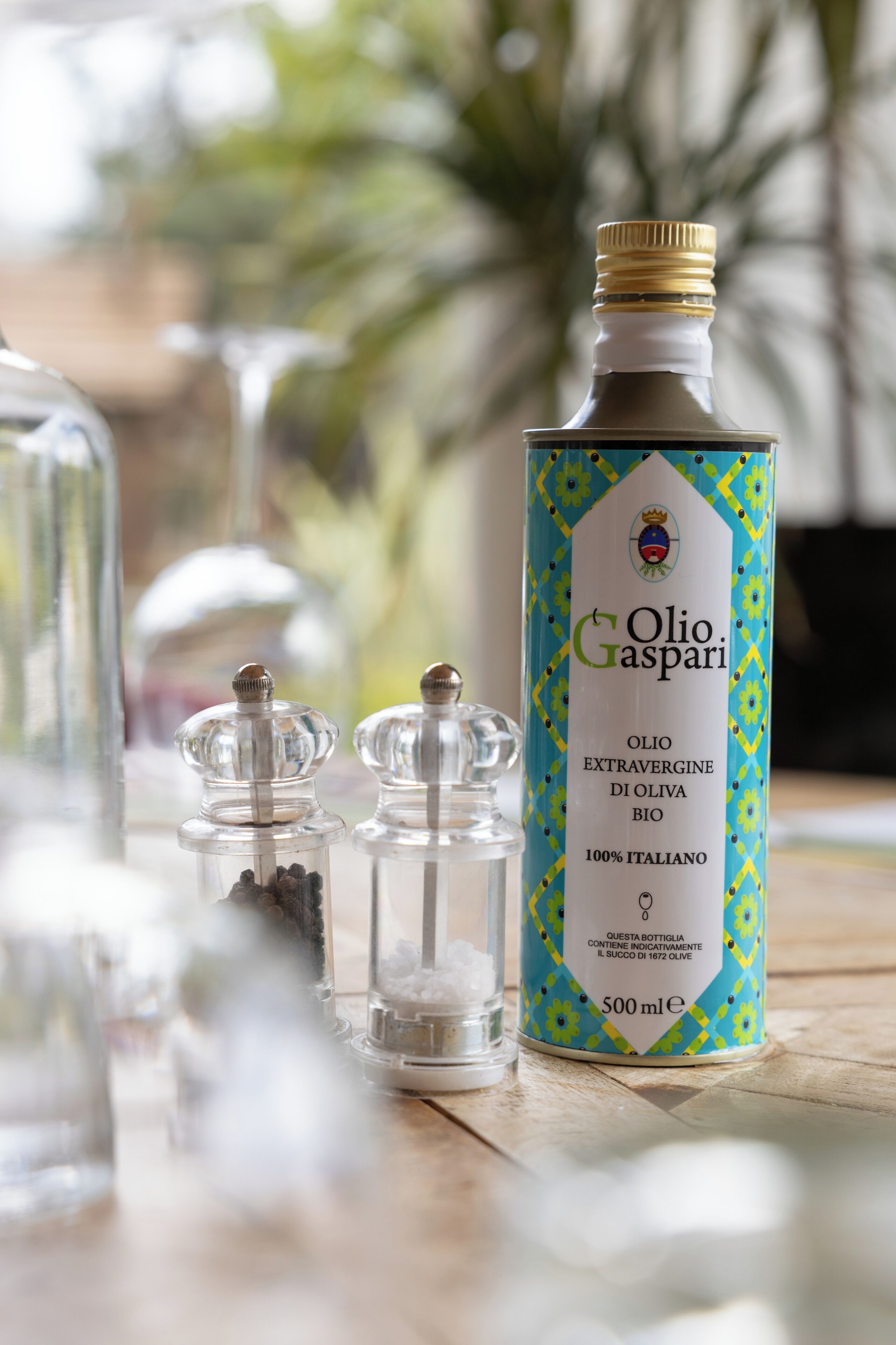 Une bouteille d’huile d’olive extra vierge Olio Gaspari, d’une capacité de 500 ml, est posée sur une surface en bois, à proximité de deux récipients en verre.