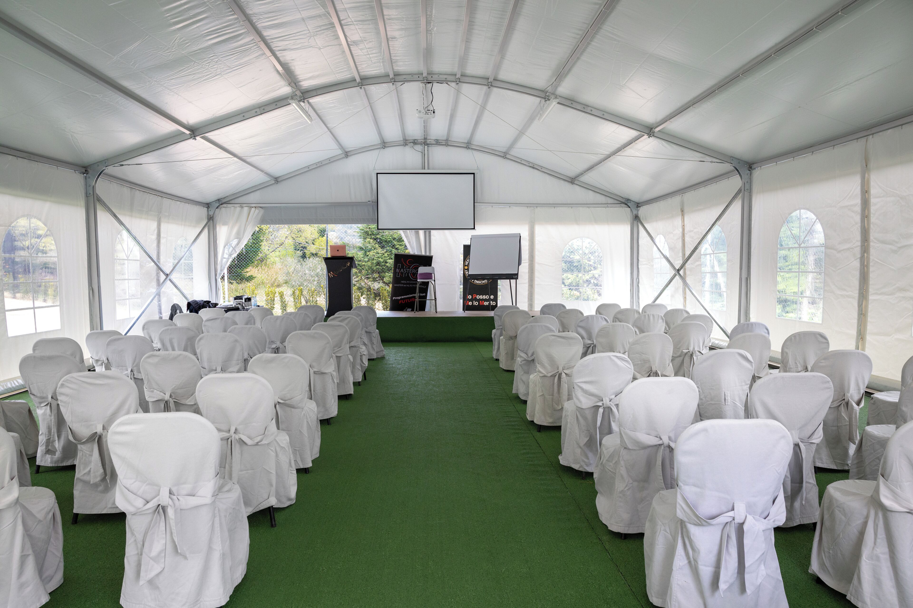 A tented event space with rows of chairs covered in white cloth, a stage with a screen, and a green carpet.