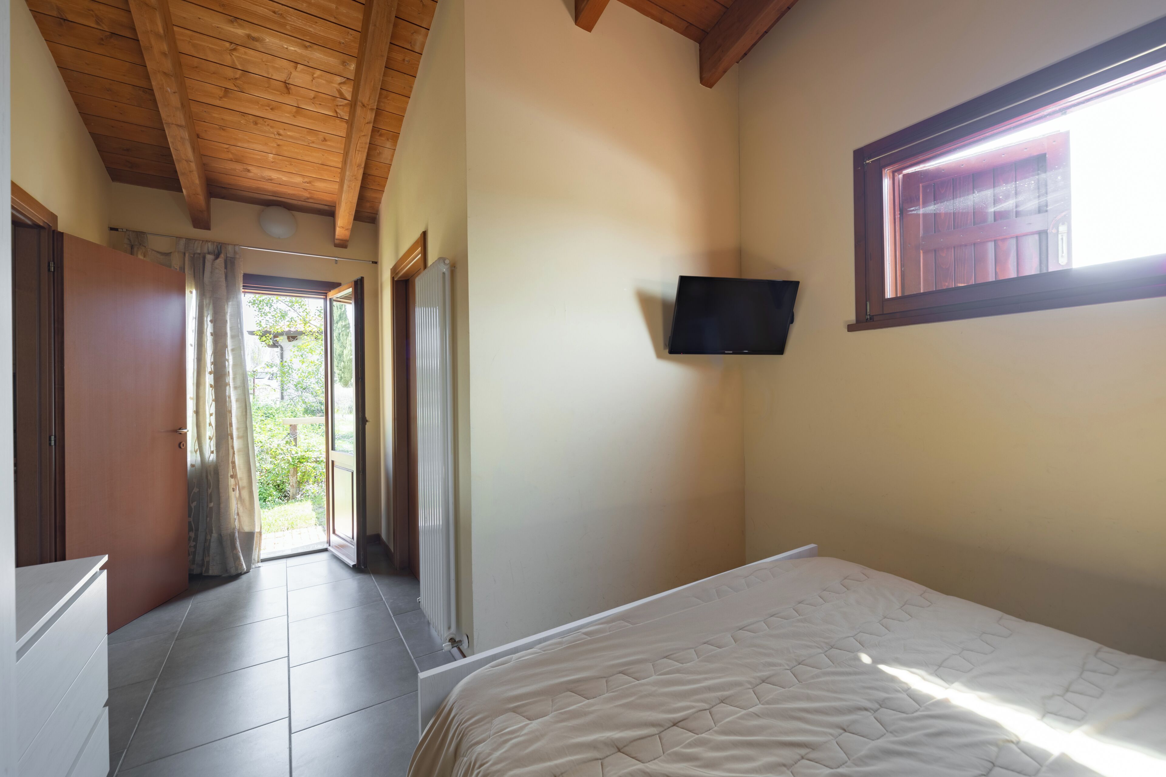 A bedroom with a bed, a television mounted on the wall, a window, and a door leading to a balcony.