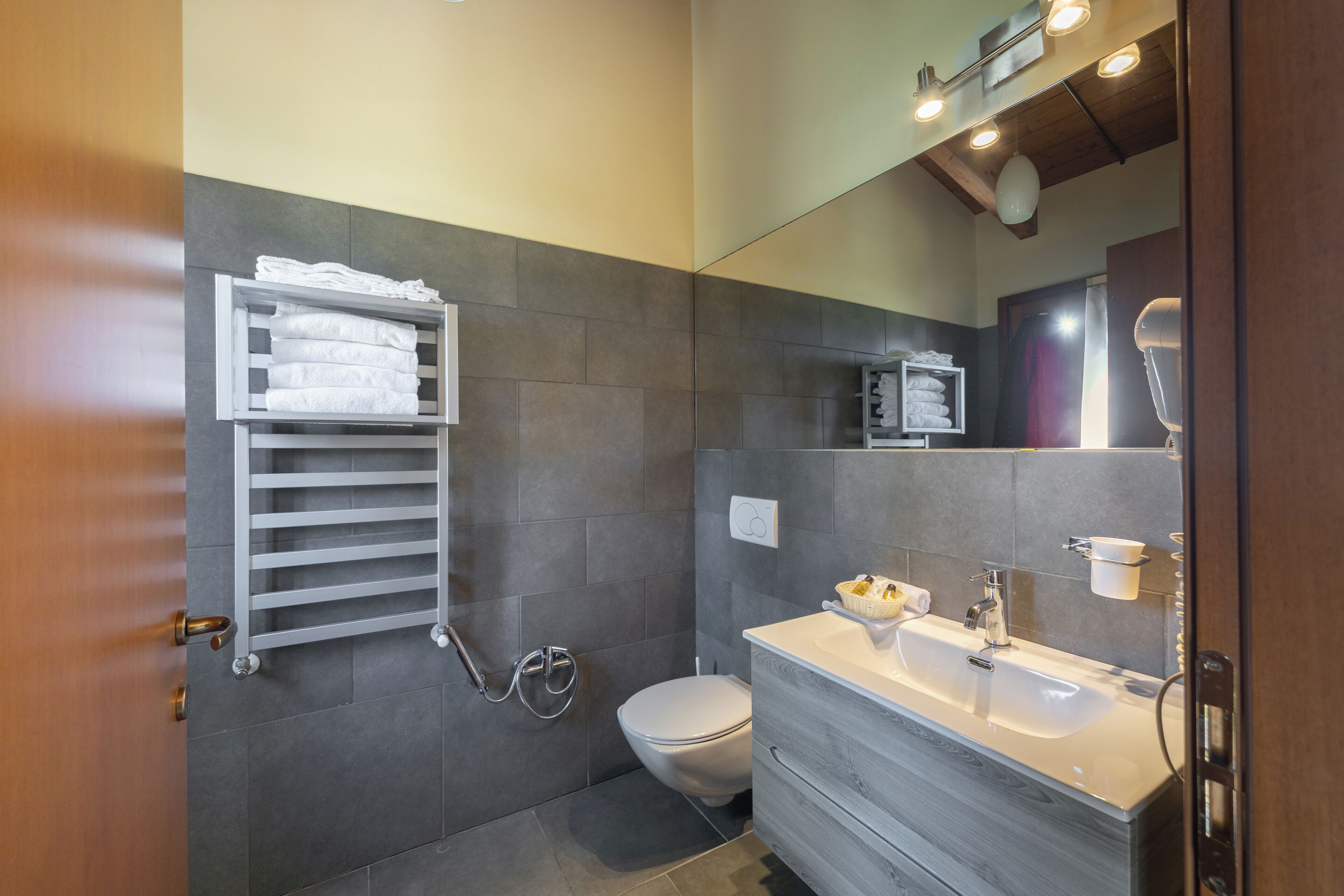 A modern bathroom with a grey tiled wall, a white toilet, a white sink with a wooden vanity, a towel rack, and a mirror with lights above.