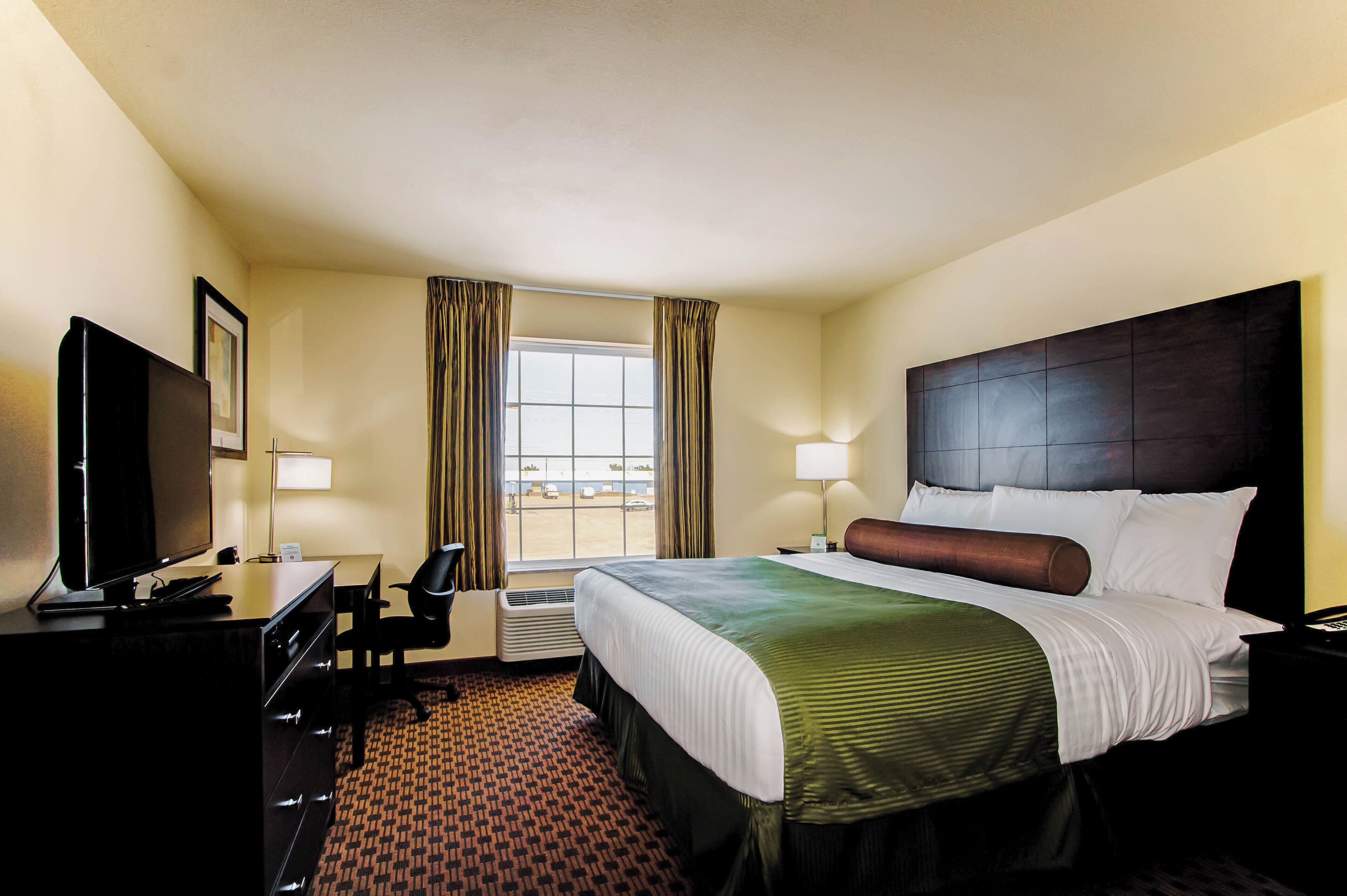 A hotel room with a bed, a desk with a computer, a TV, a window with curtains, and a nightstand with a lamp.