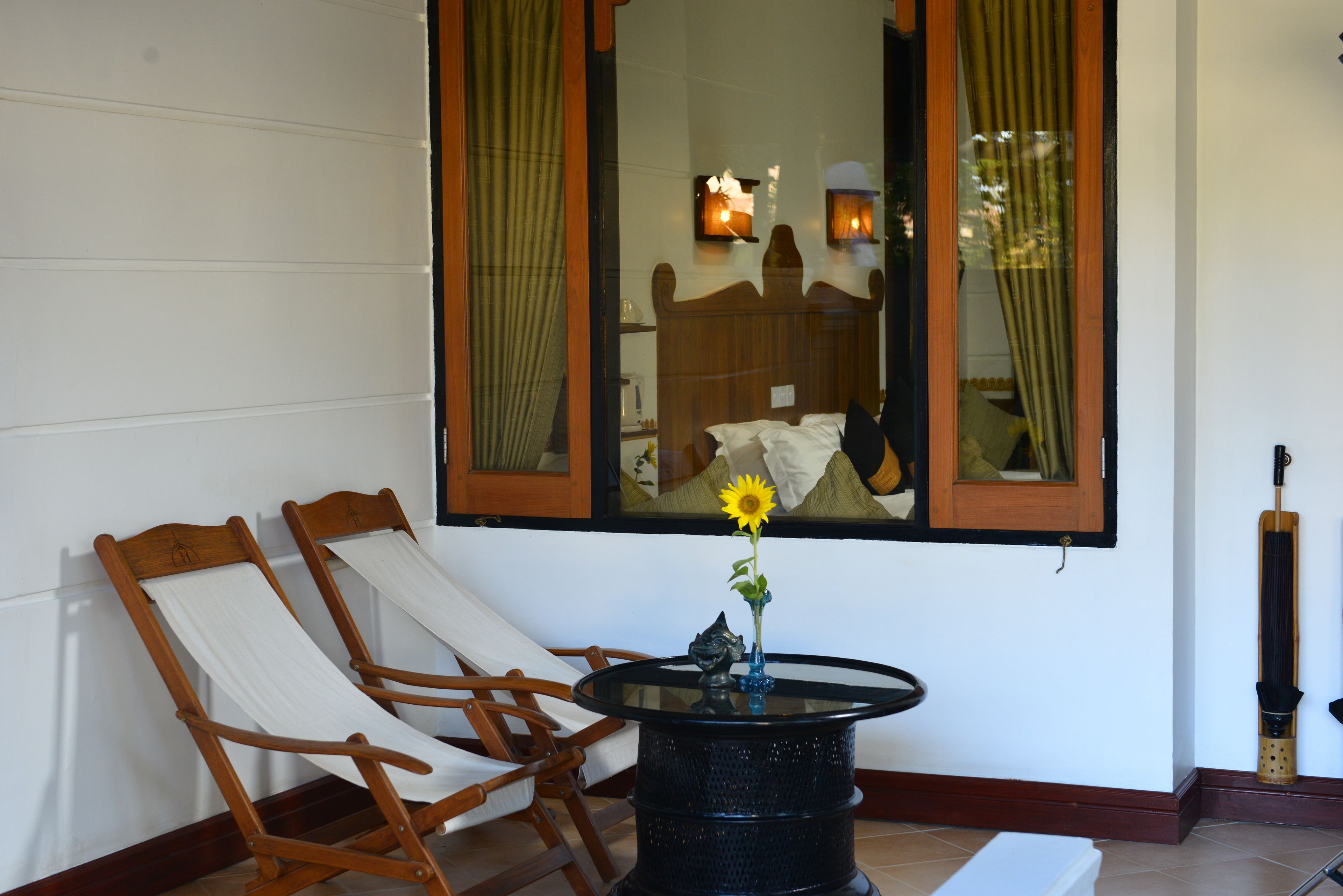 A room with two wooden chairs, a round table with a vase of flowers, and a large mirror reflecting a bed and curtains.