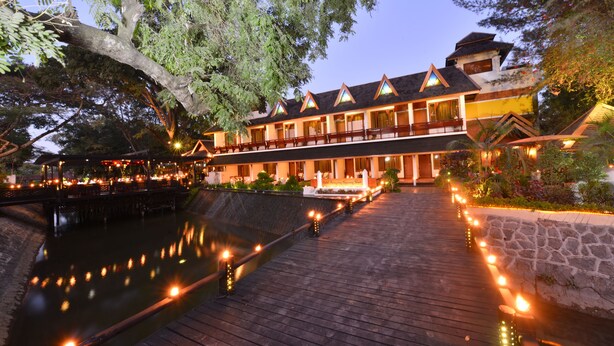 A traditional-style hotel with a wooden deck overlooking a calm body of water, surrounded by lush greenery.