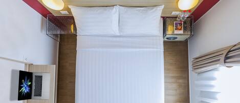 A neatly made bed with white linens, a laptop on a wooden nightstand, and a bedside table with a lamp and a bottle.