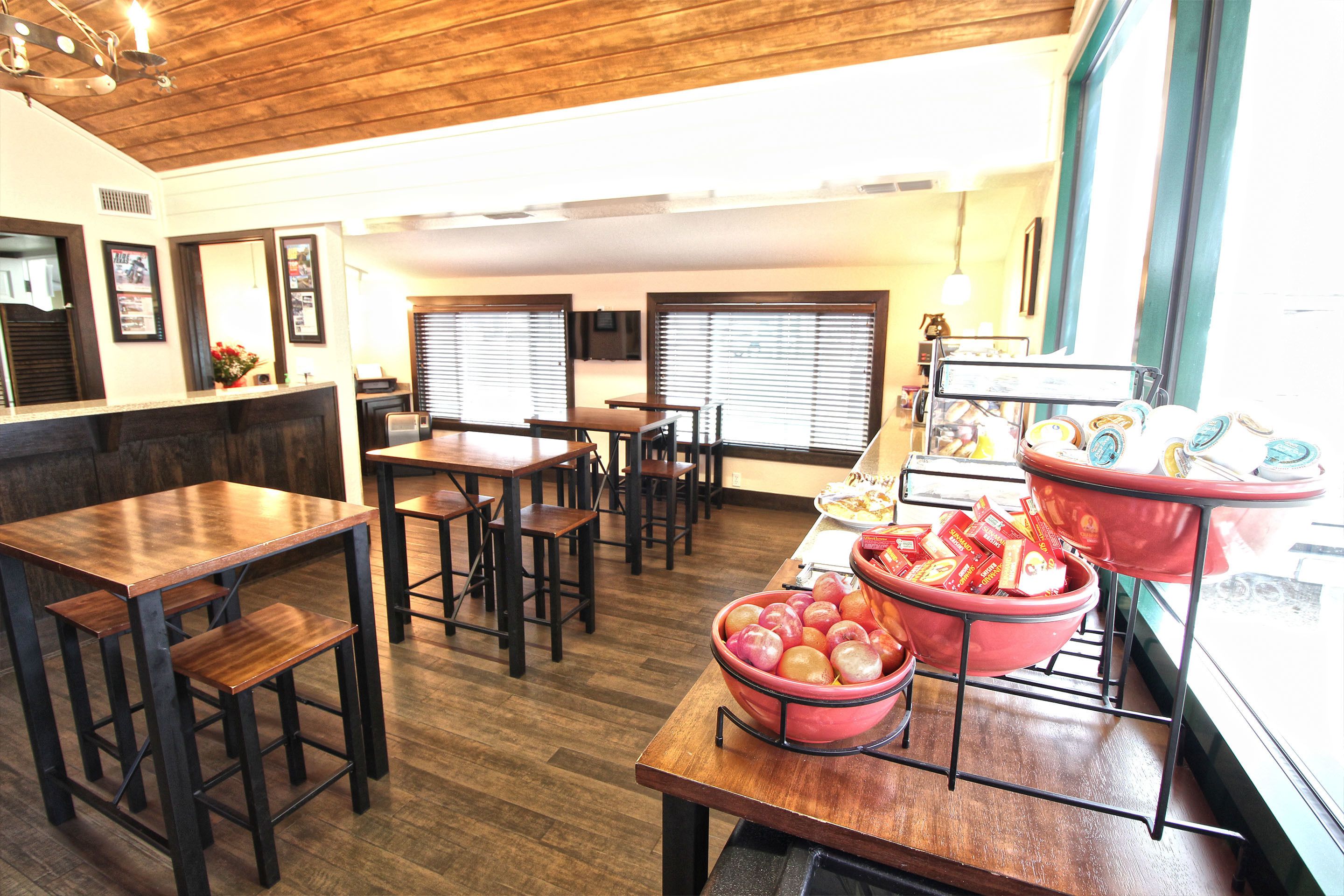A restaurant with wooden tables and chairs, a counter with food items, and large windows with blinds.