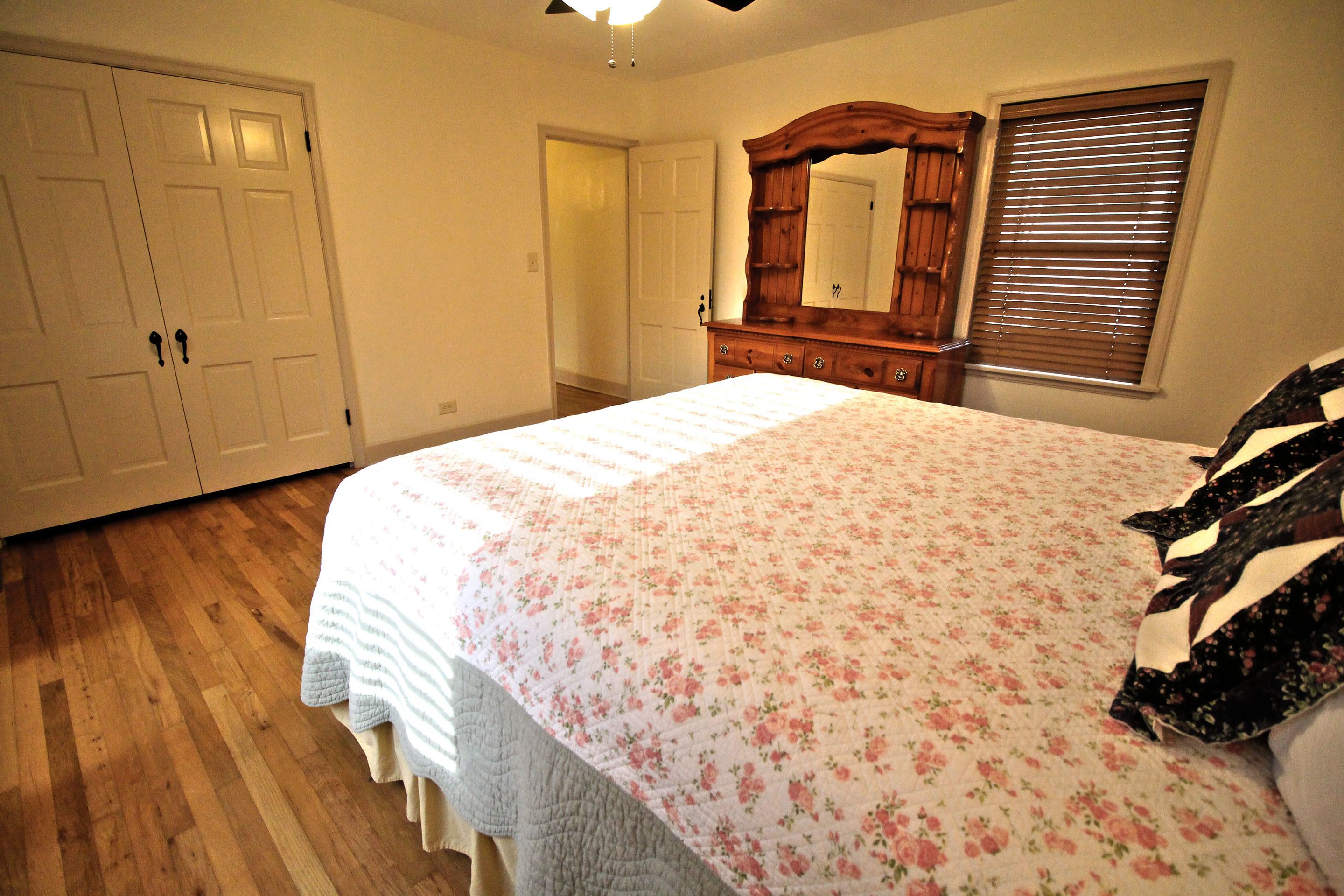 A bedroom with a large bed, a wooden dresser, a mirror, and a window with blinds.