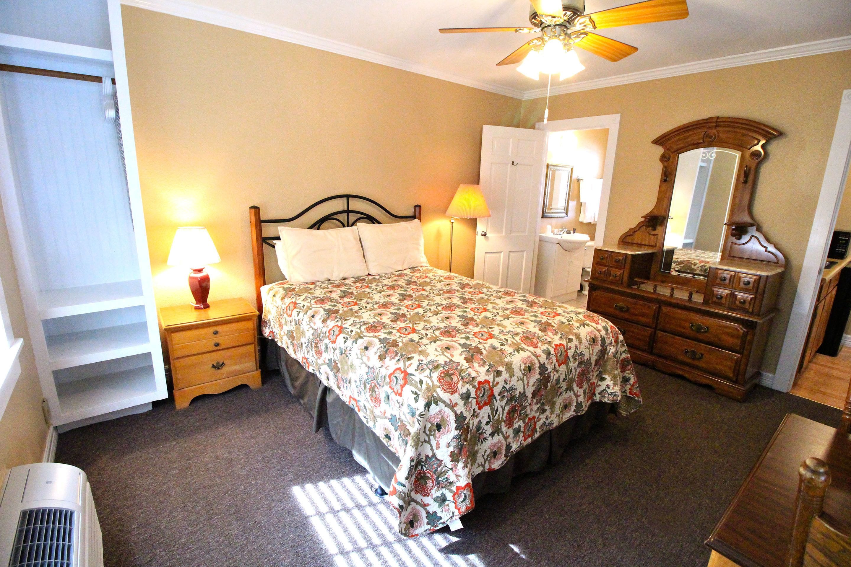 Een slaapkamer met een bed, een dressoir, een nachtkastje, een lamp, een spiegel en een plafondventilator.