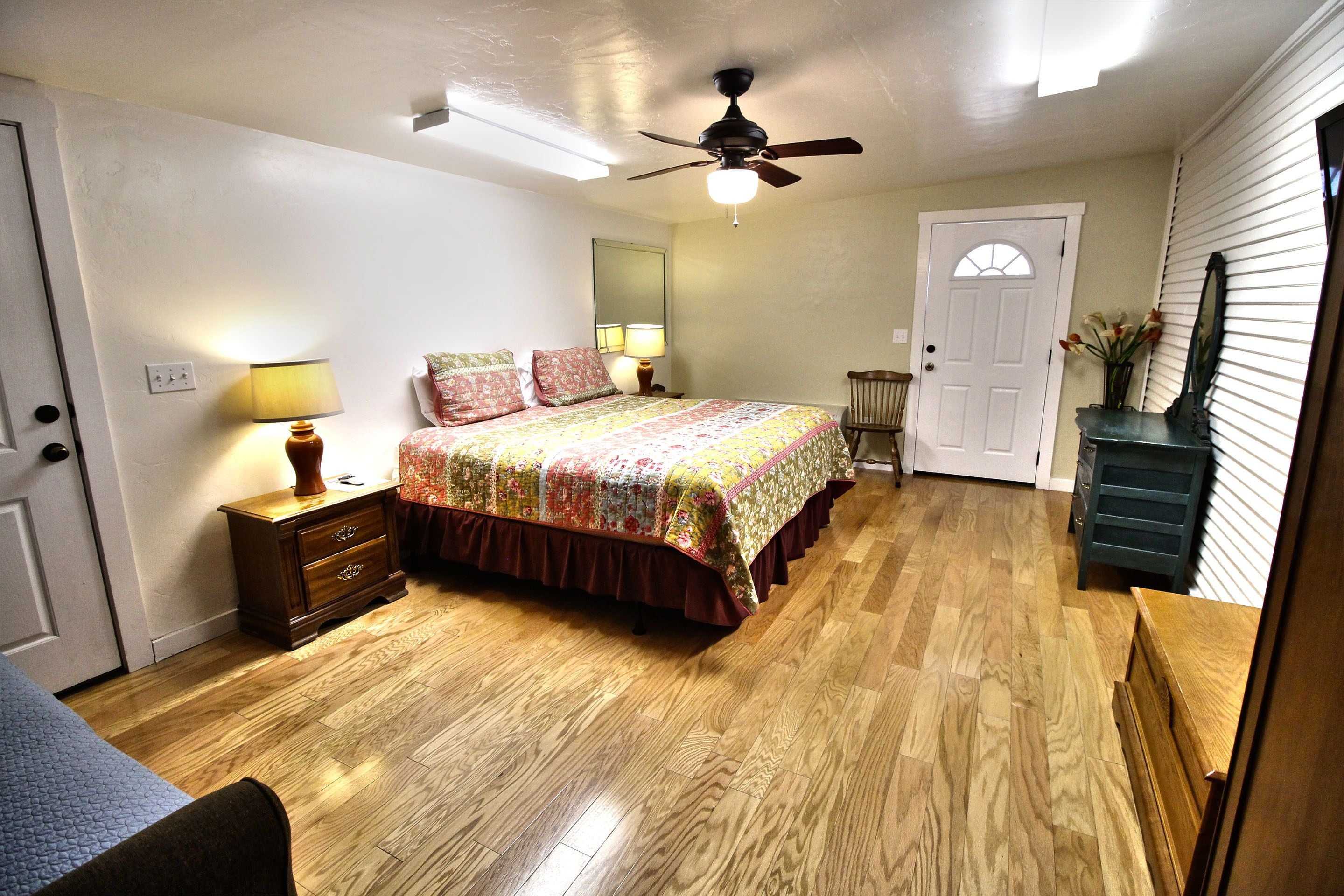 A bedroom with a wooden floor, a bed with a floral bedspread, a ceiling fan, a nightstand with a lamp, and a dresser.