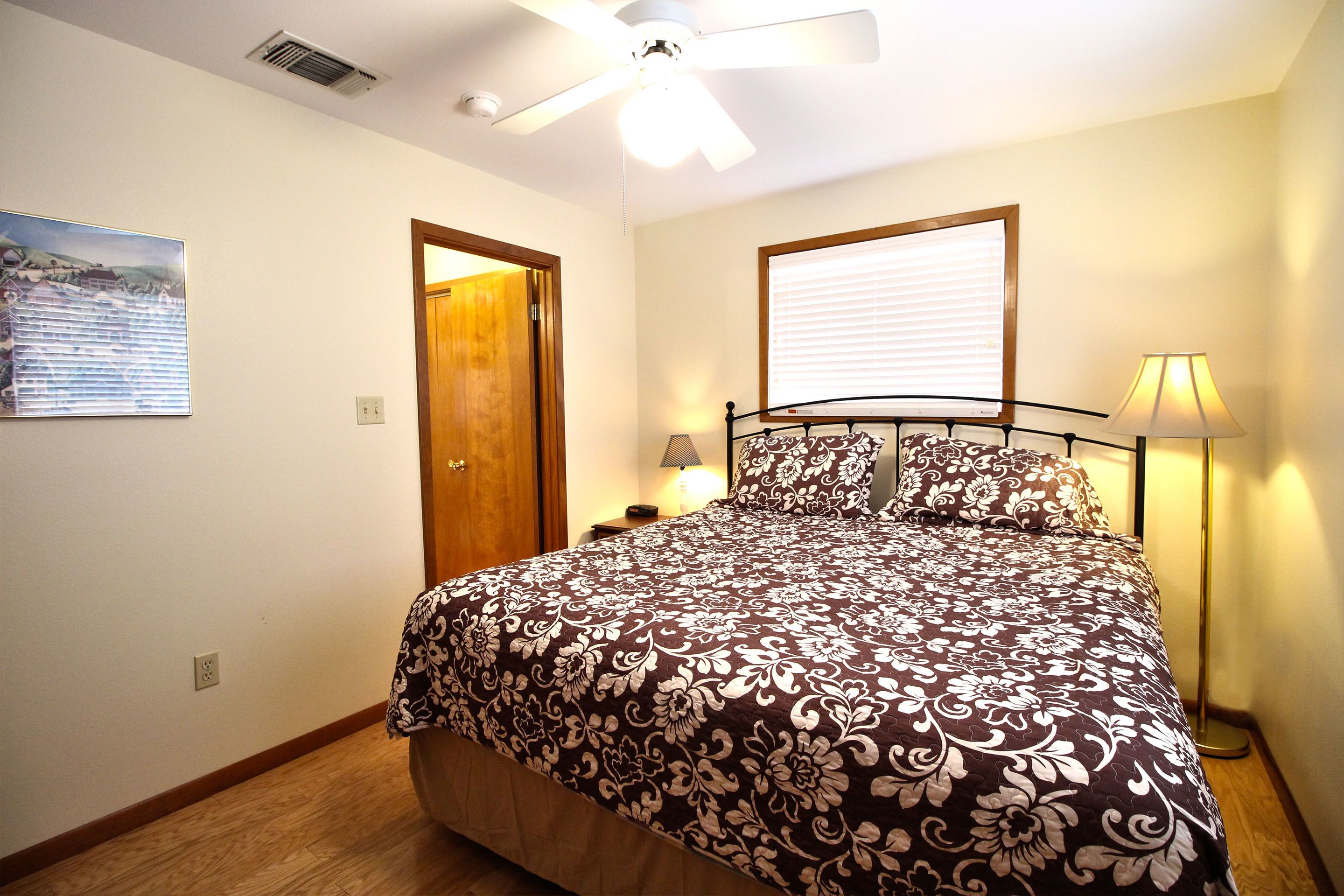 A bedroom with a bed, a ceiling fan, a window with blinds, a nightstand with a lamp, and a door.