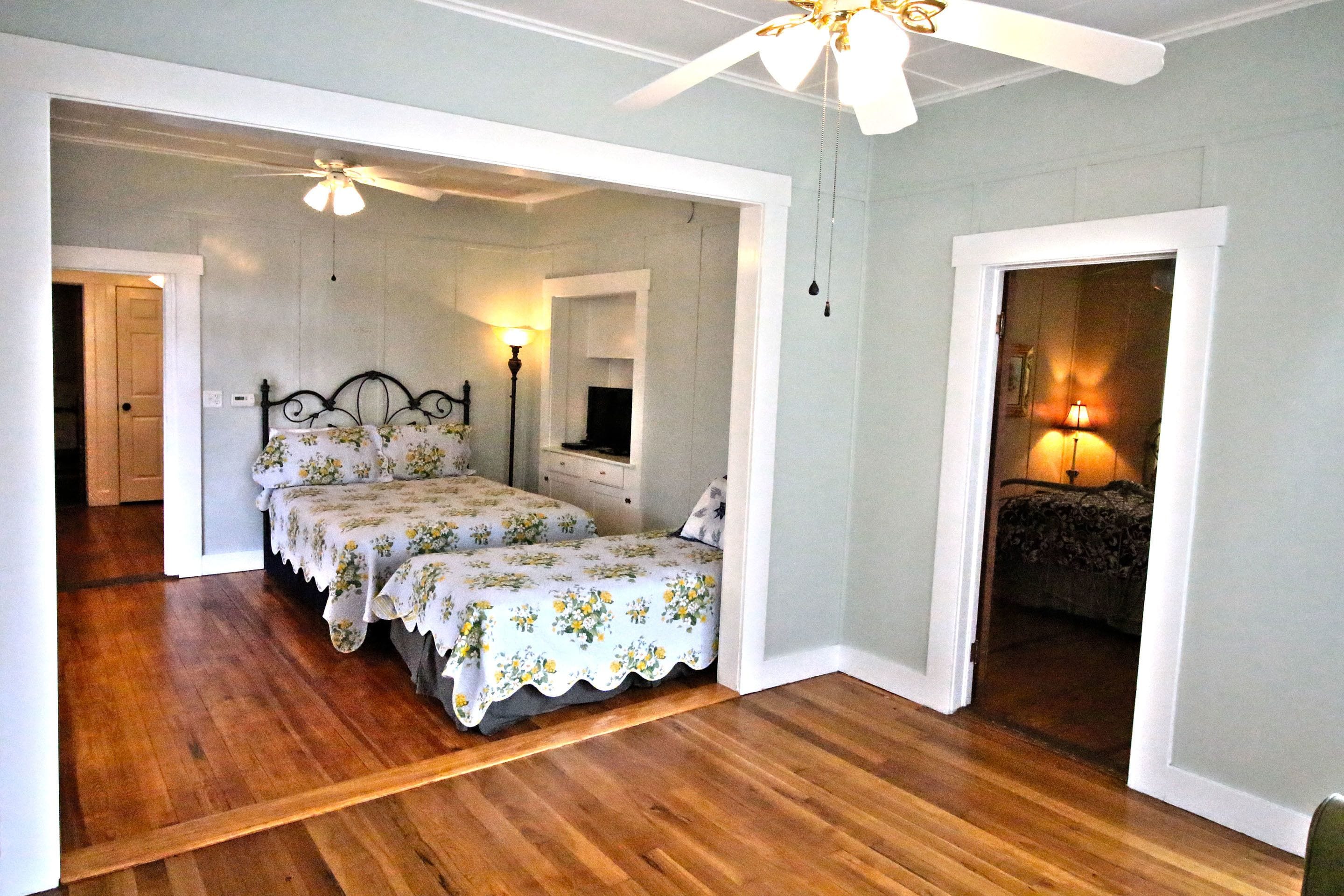 A bedroom with two beds, wooden flooring, a ceiling fan, and a nightstand with a lamp.