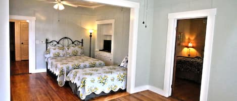A bedroom with two beds, wooden flooring, a ceiling fan, and a nightstand with a lamp.