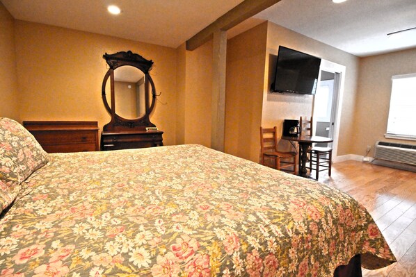 Une chambre avec une couvre-lit à motifs floraux, une commode en bois, un miroir, un téléviseur fixé au mur et un bureau avec une chaise.
