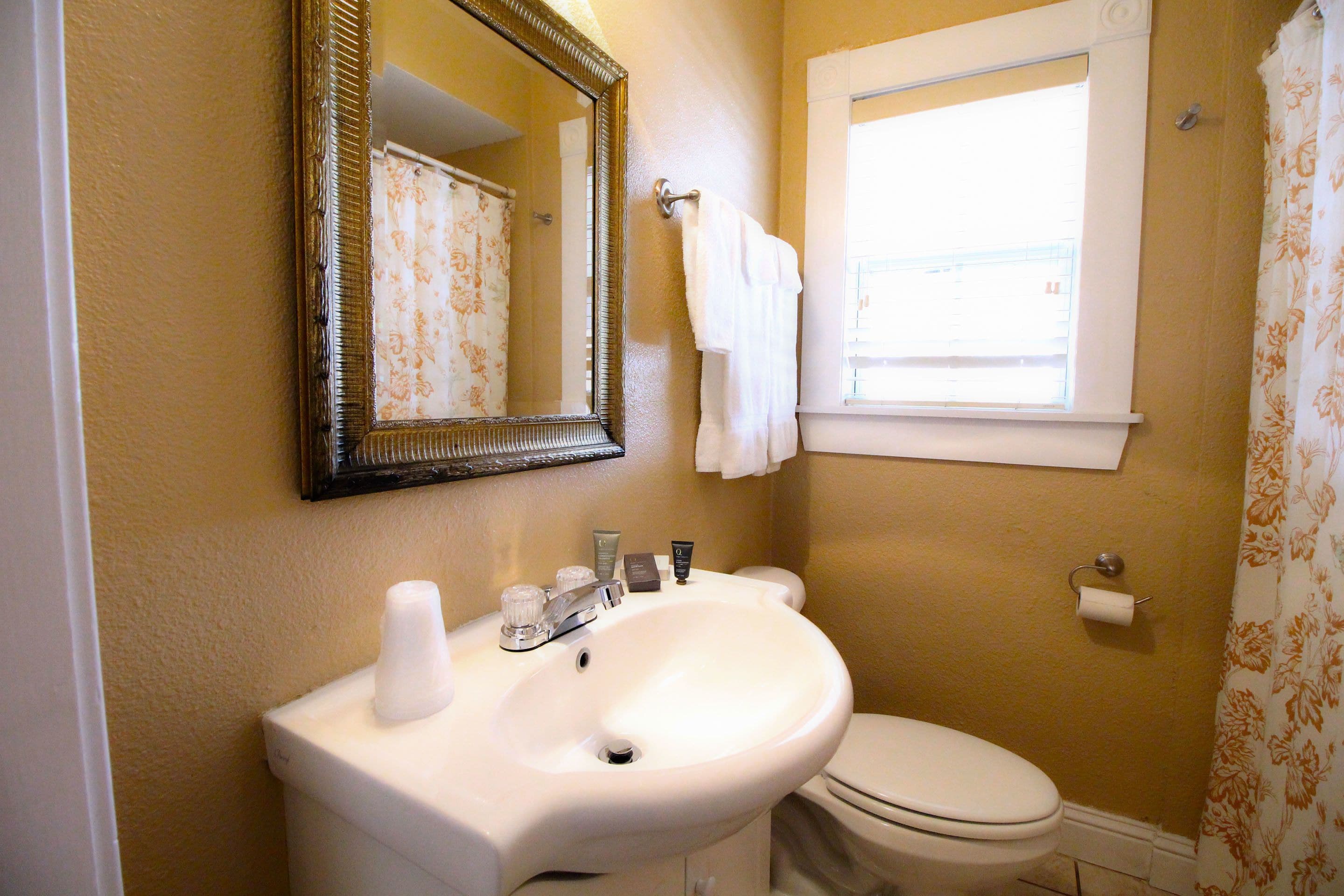 Un baño con un lavabo blanco, un inodoro, un espejo y una ventana con una toalla colgada en la pared.