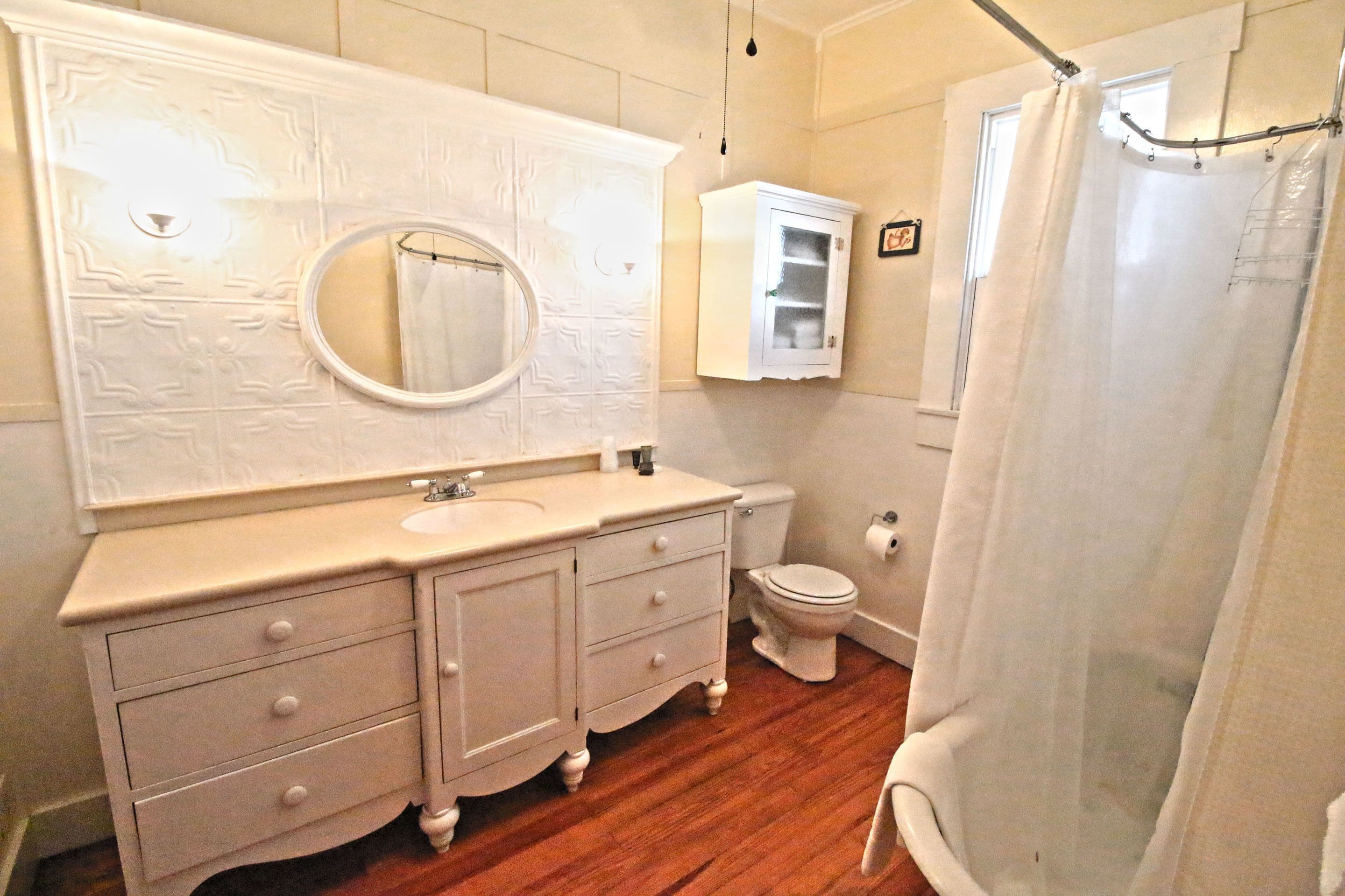 Une salle de bain avec un plancher en bois, un meuble-lavabo blanc doté de tiroirs, un miroir rond, des toilettes et un rideau de douche.