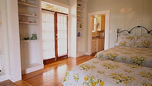 A bedroom with a wooden floor, a bed with floral bedding, a wardrobe, and a kitchen area visible through an open door.