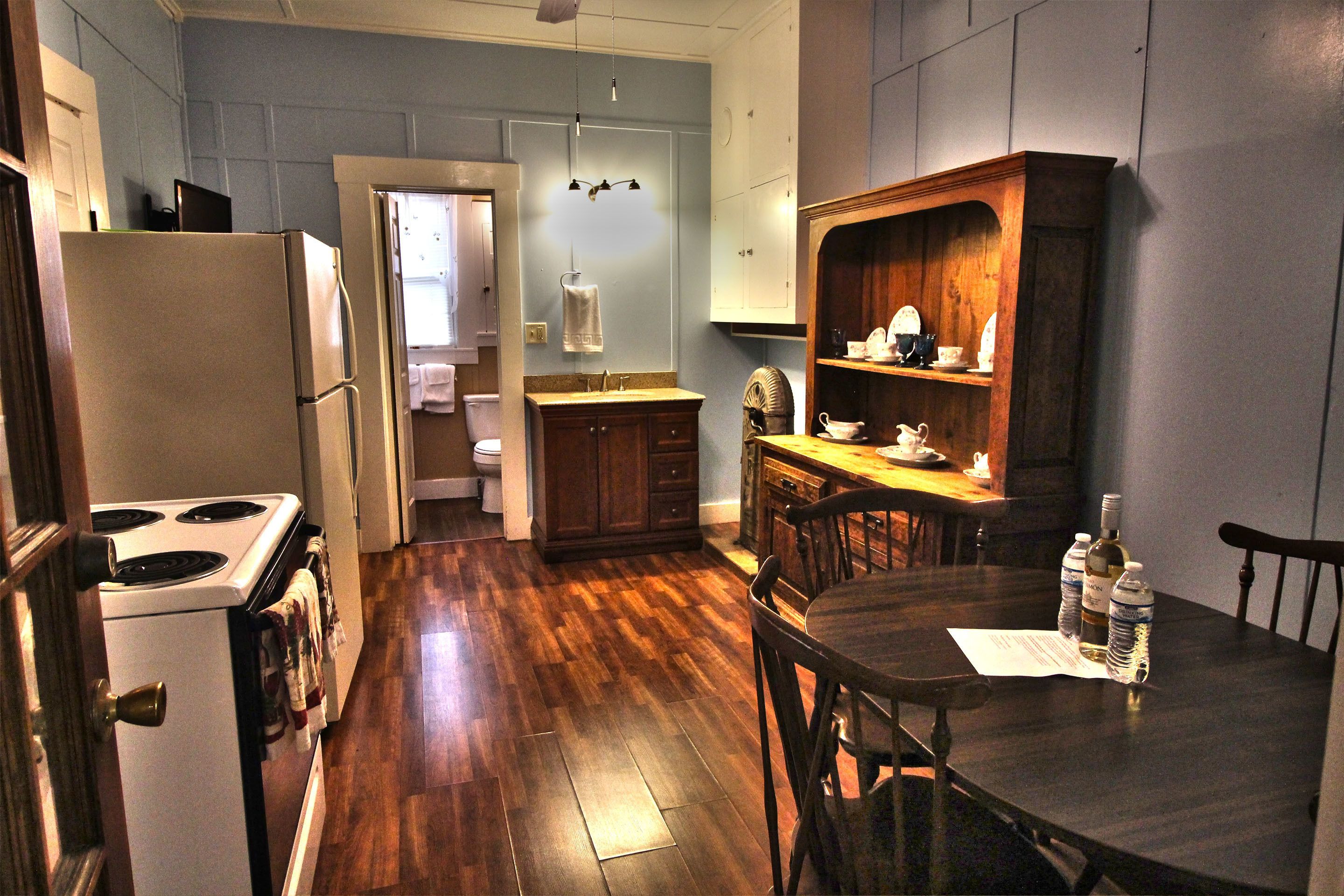 A kitchen with wooden flooring, a dining table with chairs, a refrigerator, a stove, and a cabinet with dishes.